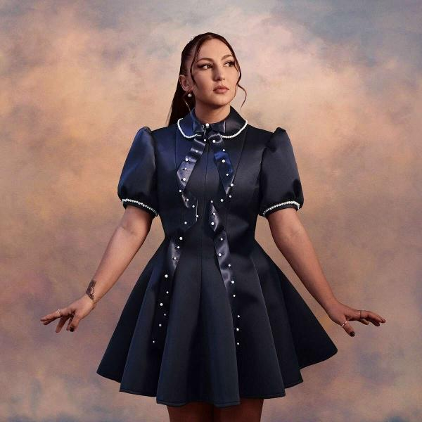 Een vrouw in een marineblauwe jurk met witte kraag en parelaccenten staat met uitgestrekte armen. De achtergrond is een bewolkte lucht.