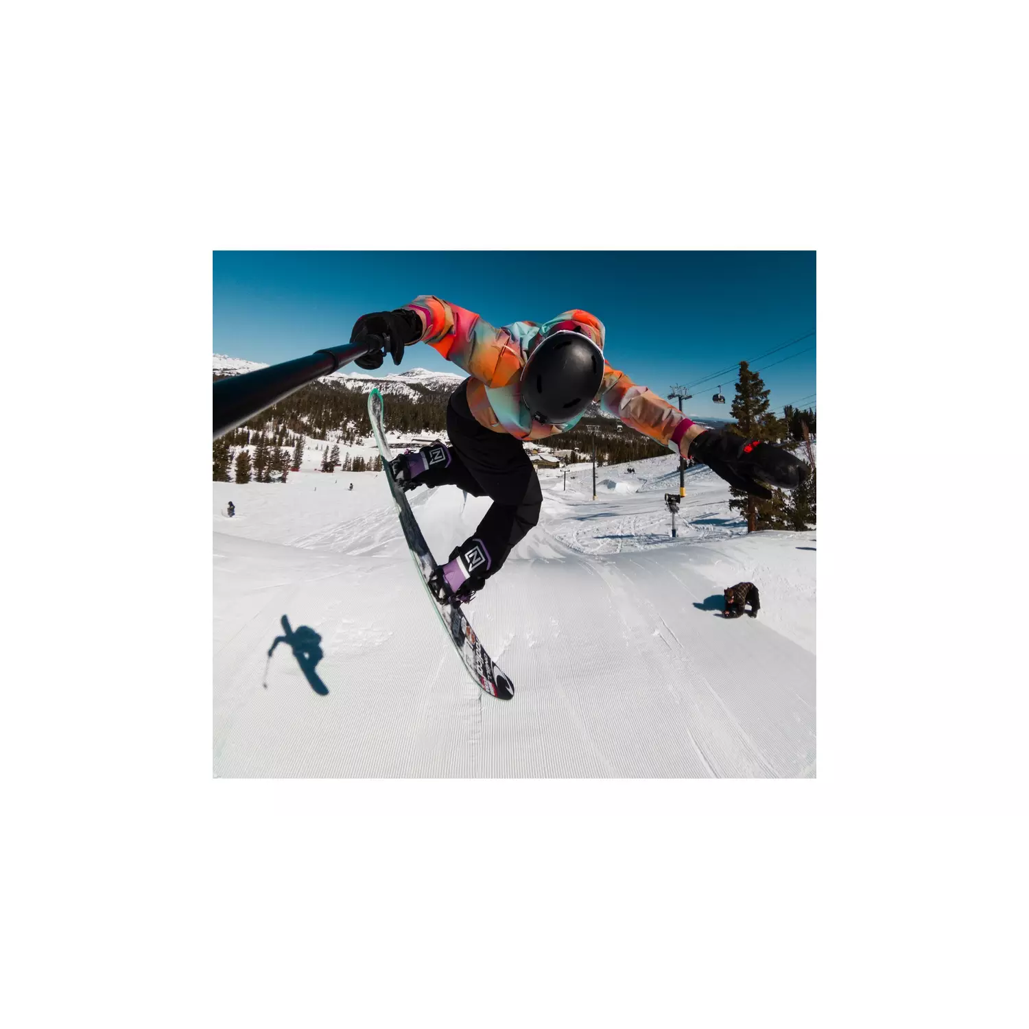 Snowboarder in de lucht, met een selfiestick. Draagt een kleurrijke jas, zwarte helm en broek. Besneeuwde berg op de achtergrond.