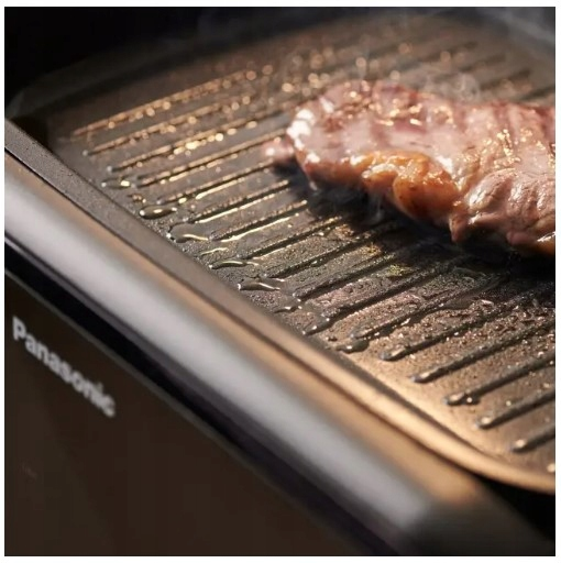Kawałek mięsa jest na gorącym, czarnym grillu z olejem. Para unosi się. Widoczne jest słowo 'Panasonic'.