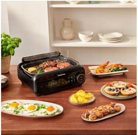 Czarny grill Panasonic z jedzeniem, w tym szaszłykami, burgerami i różnymi daniami na talerzach. Tło: półka z naczyniami.