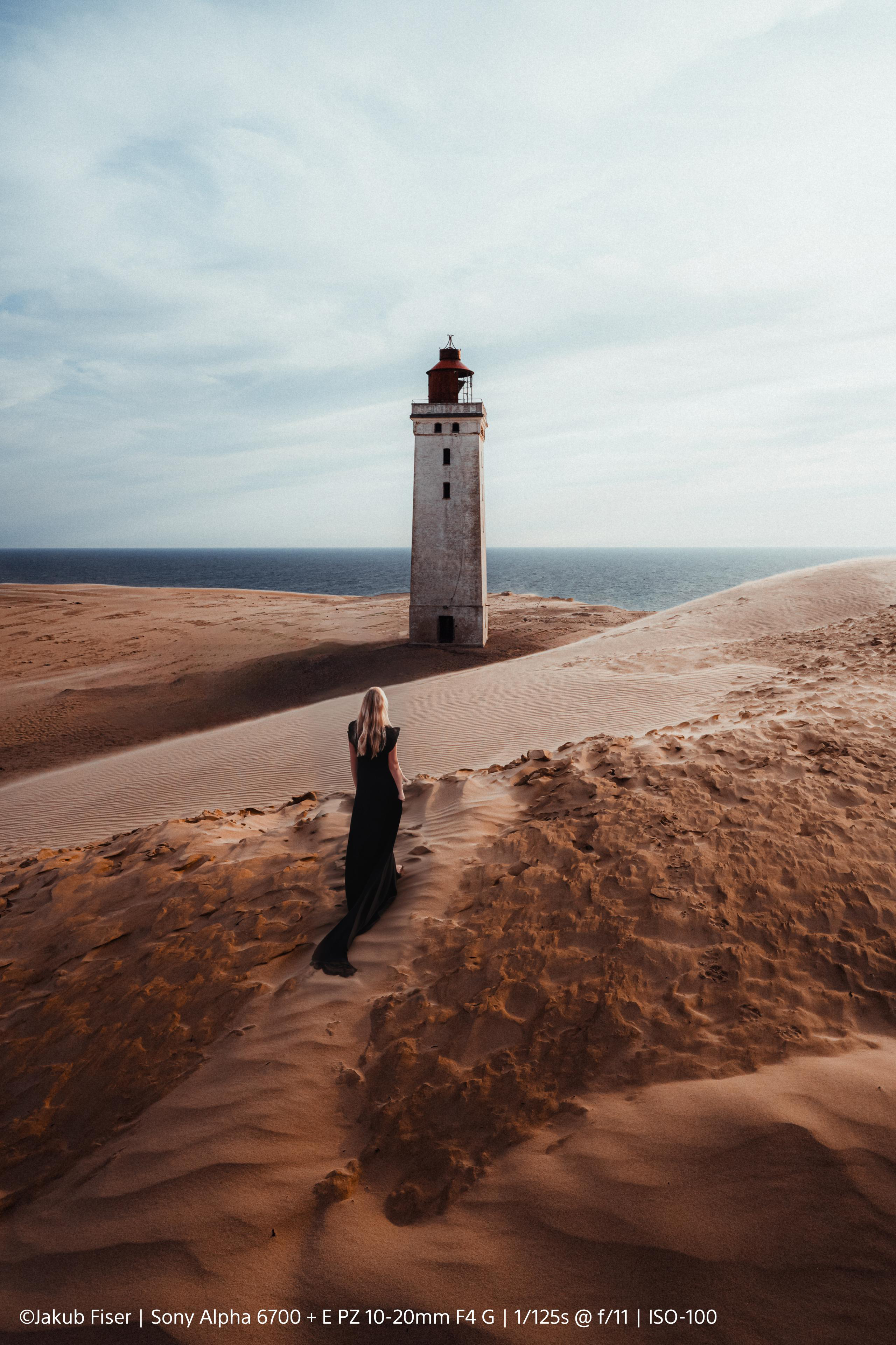 Frau in schwarzem Kleid geht auf einen Leuchtturm auf Sanddünen zu, Meer und Himmel im Hintergrund.