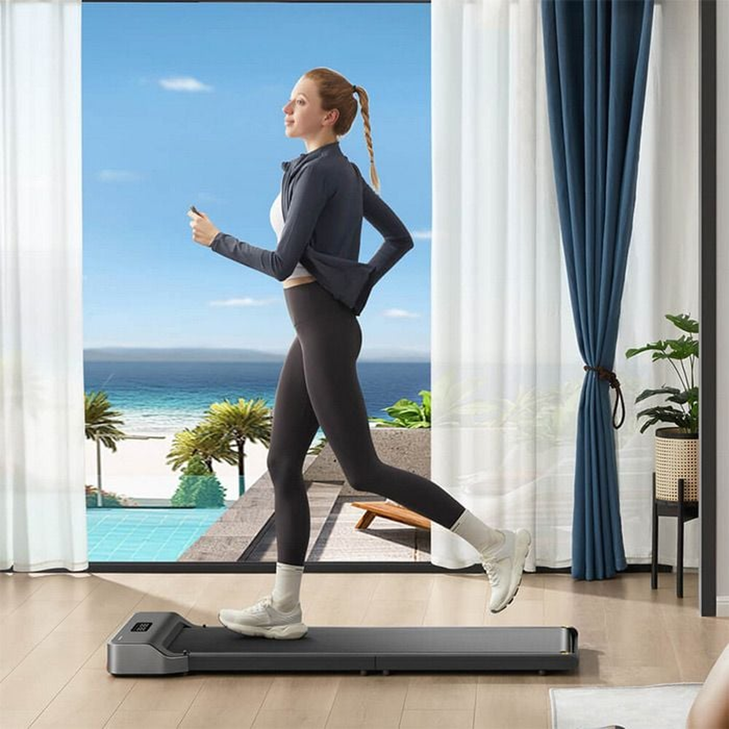 A woman exercises on a treadmill by a window overlooking the ocean. She wears a dark grey jacket, leggings, and white sneakers.
