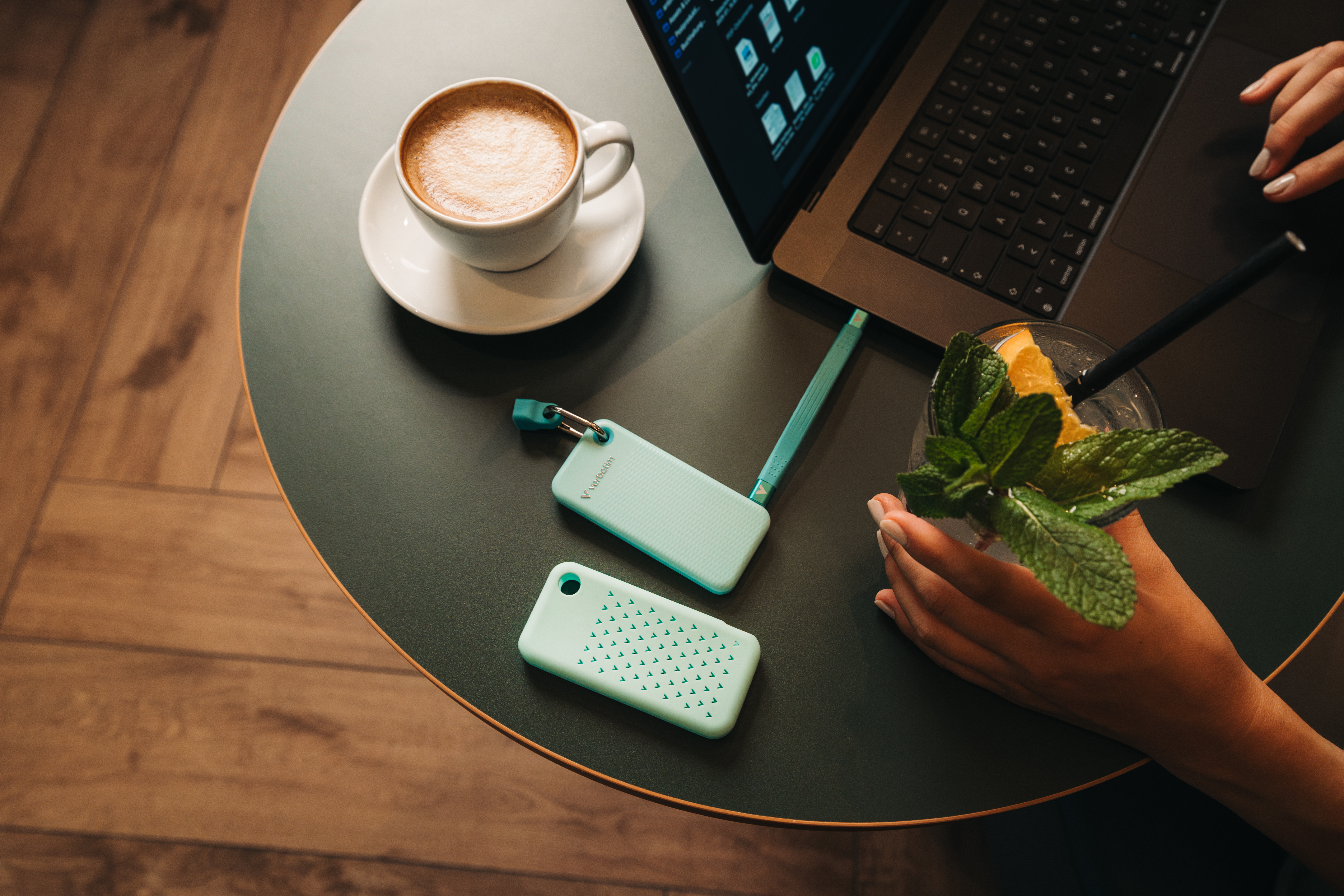 Tisch mit Kaffee, Laptop, Getränk und zwei mintgrünen Geräten. Hände einer Frau sichtbar. Die Szene ist gut beleuchtet, mit moderner Ästhetik.