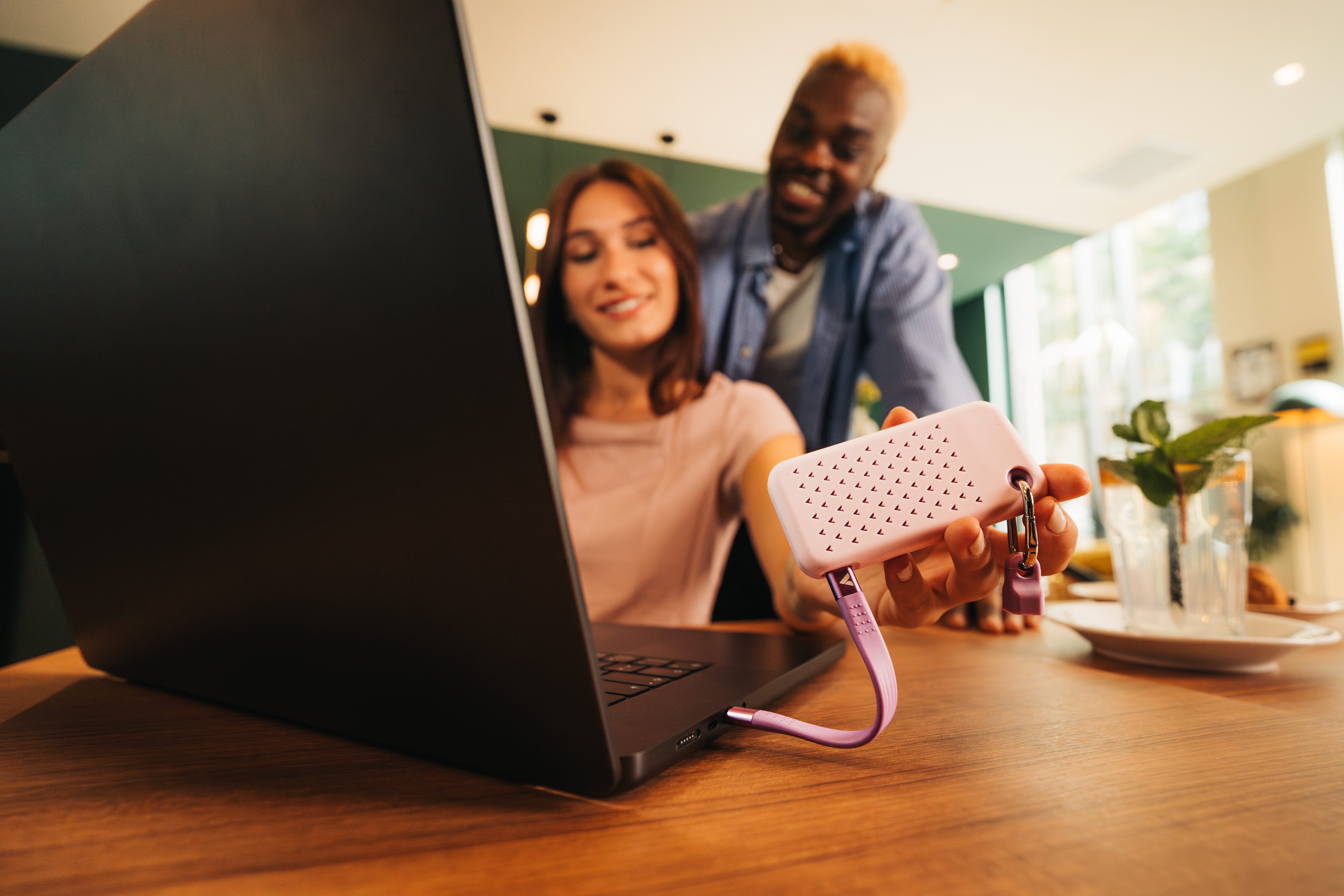 Eine Frau und ein Mann schauen auf einen Laptop. Ein rosa Gerät ist an den Laptop angeschlossen. Es ist in einem Café.