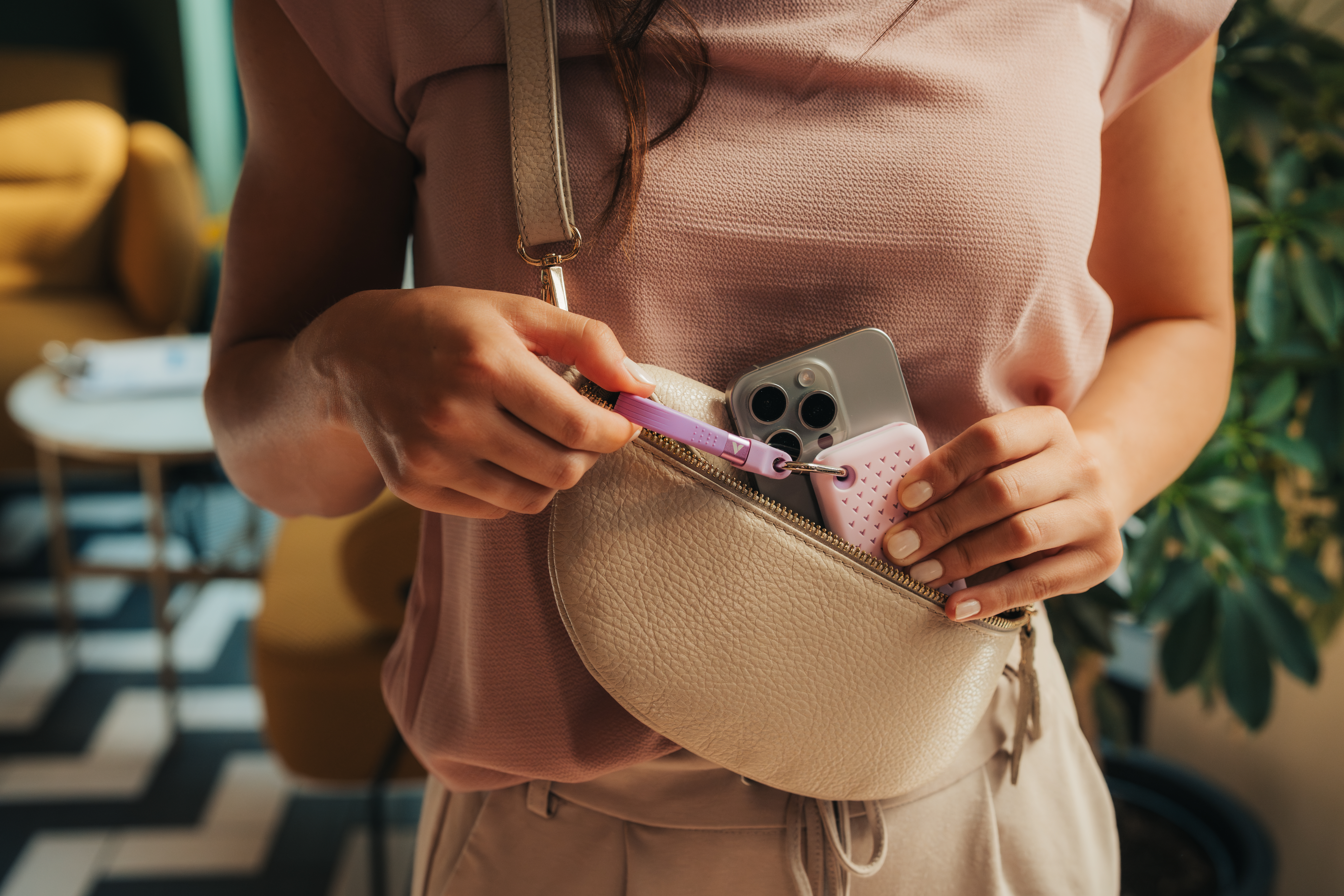Eine Person, die eine hellrosa Handtasche mit Handy, rosa Hülle und lila Schlüsselanhänger hält. Die Person trägt ein rosa Oberteil.