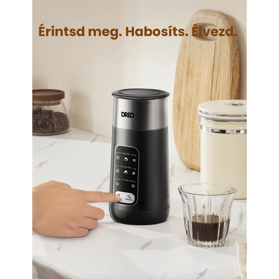 Egy kéz megnyom egy gombot egy fekete tejhabosítón. Egy pohár kávé van mellette. Más üvegek és egy vágódeszka a háttérben.