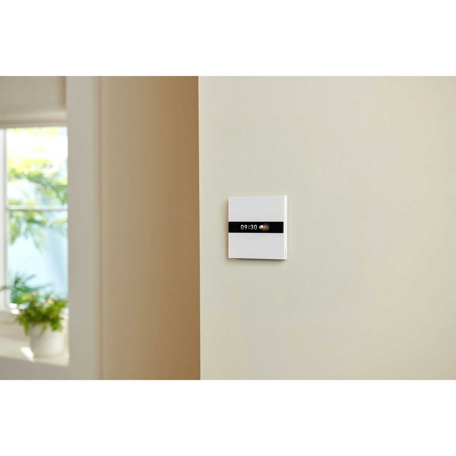 A white wall-mounted smart home device displays time and weather. A window with plants is in the background.