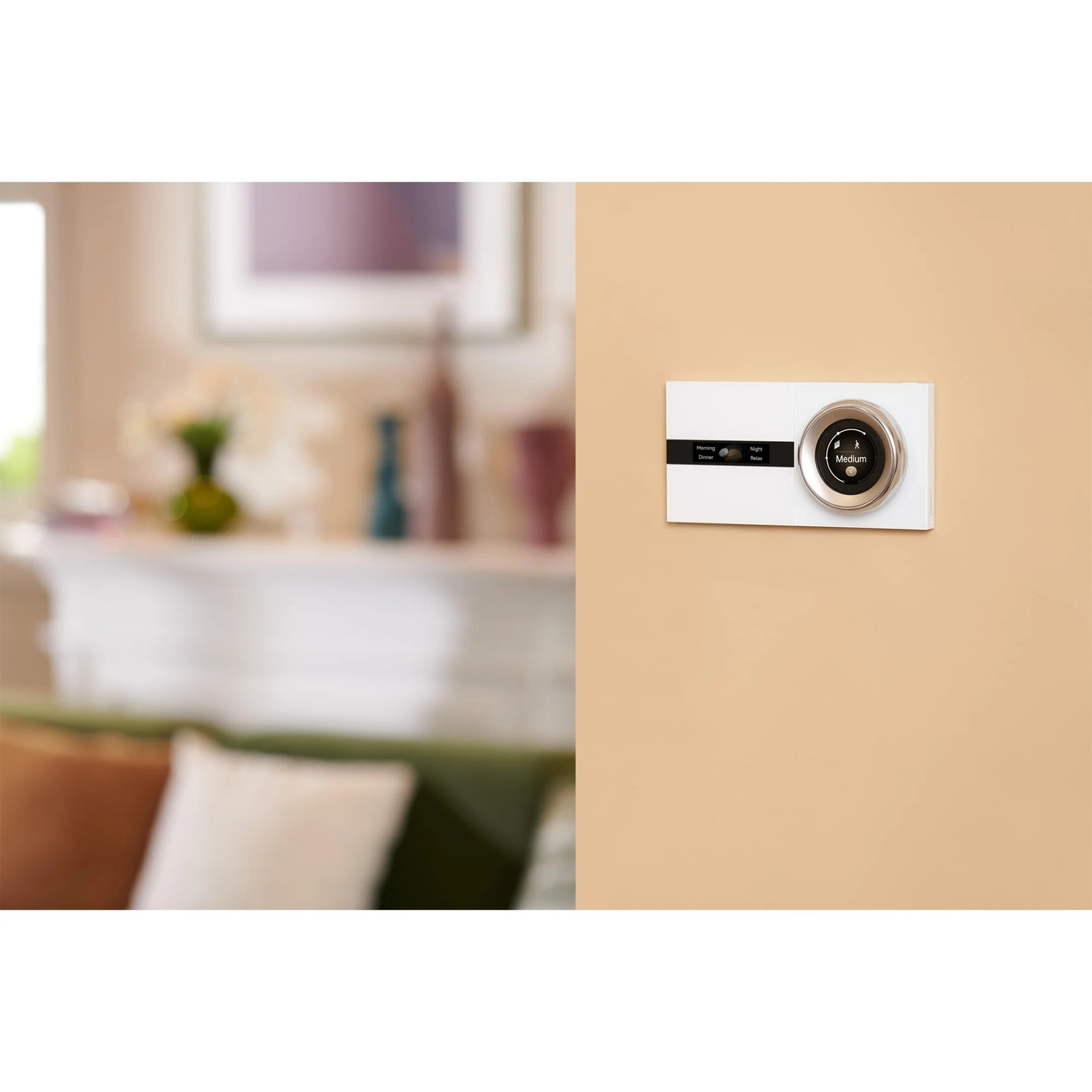 A thermostat on a beige wall. A blurred living room is in the background. The thermostat has a round dial and a digital display.