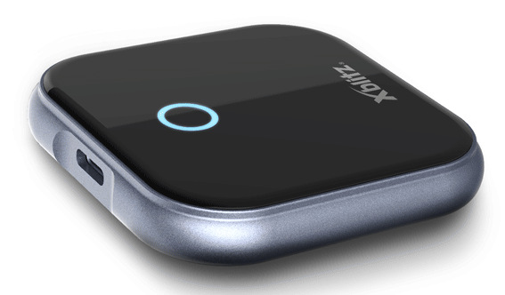 Czarno-szare urządzenie Xblitz. Ma niebieskie światło i srebrną ramkę. Urządzenie leży na białej powierzchni.