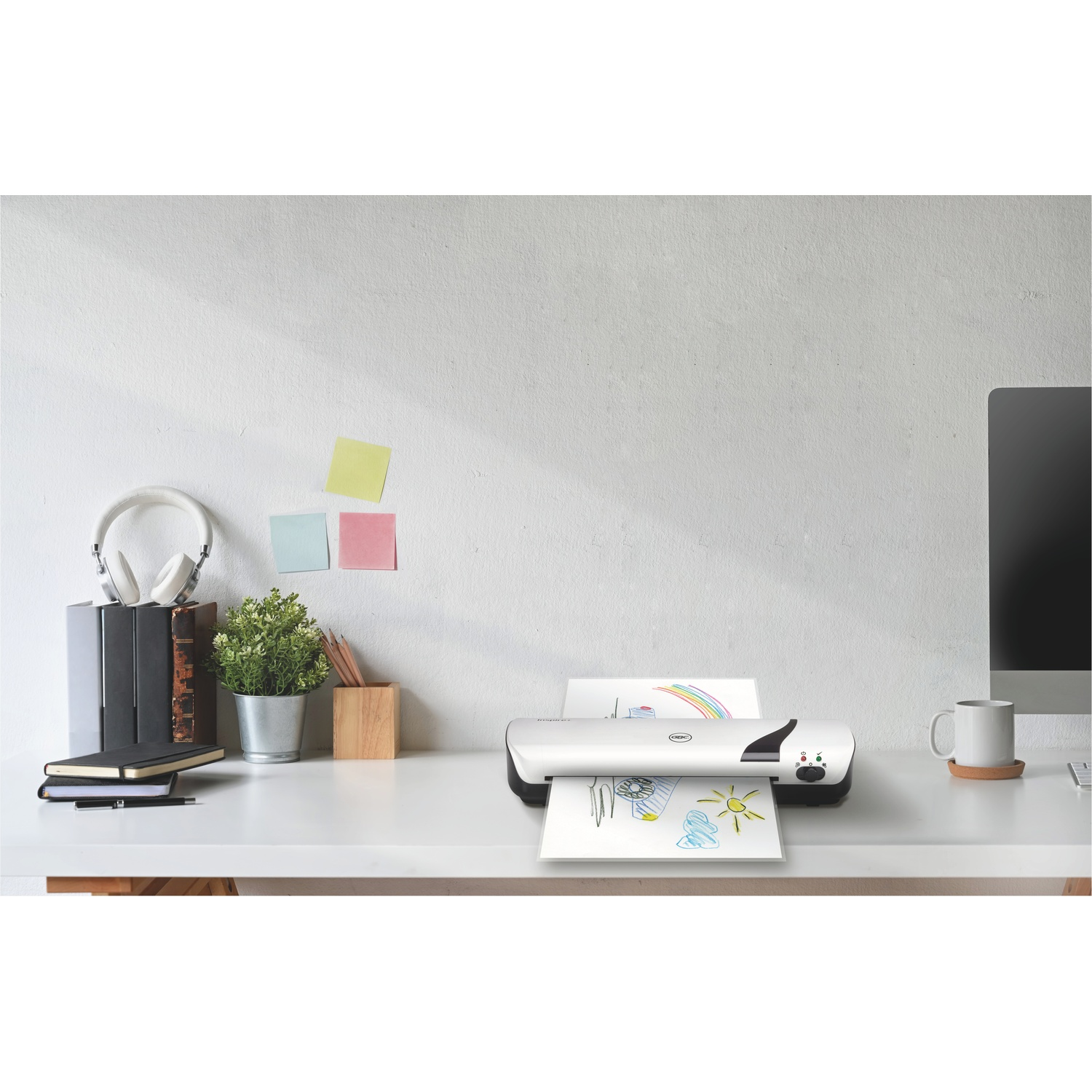 Una scrivania bianca con una plastificatrice, un monitor, una tazza, cuffie e altri oggetti per l'ufficio. Un disegno esce dalla plastificatrice.