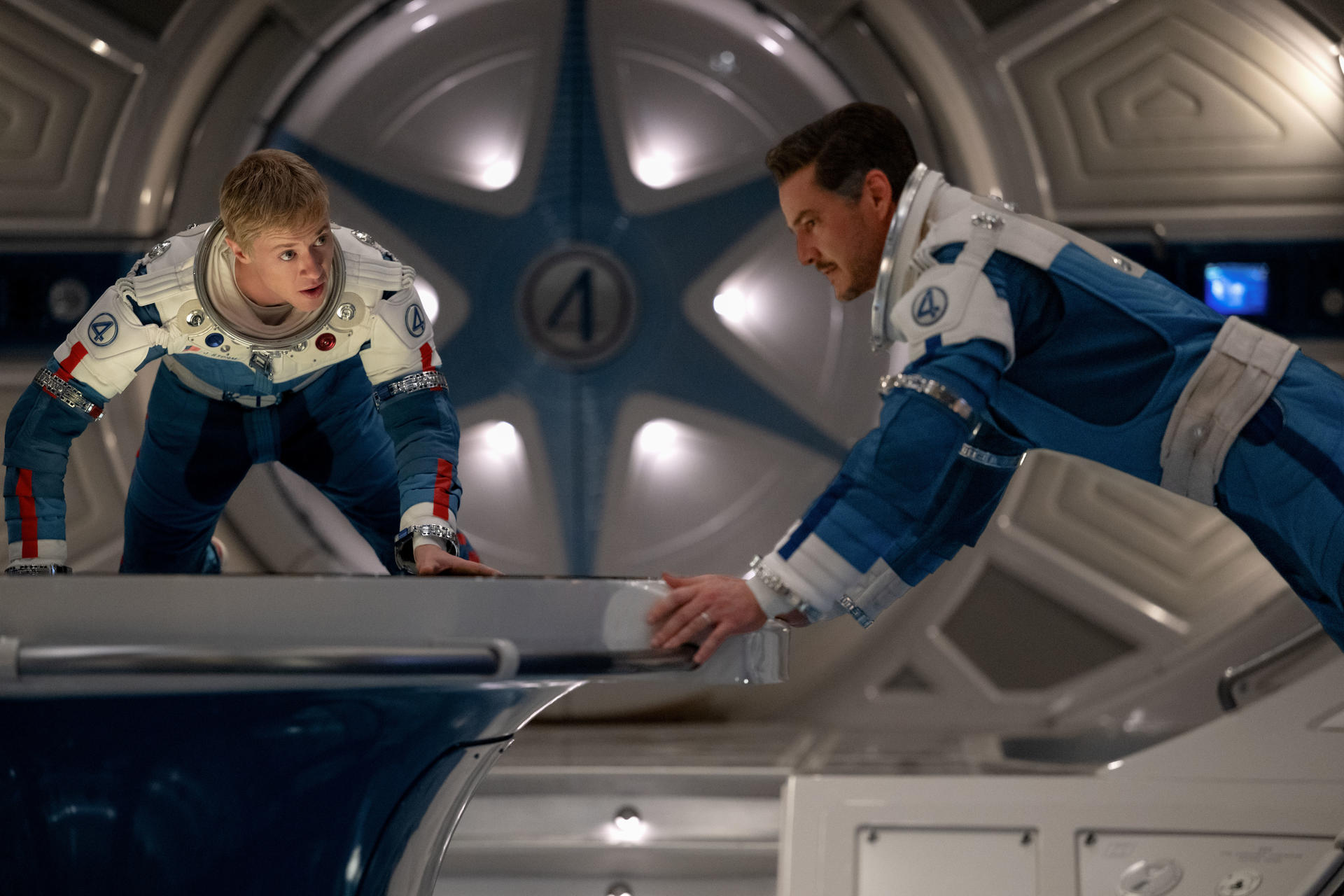 Zwei Astronauten in blau-weißen Raumanzügen beugen sich über eine Oberfläche. Einer blickt nach oben, der andere nach unten. Eine Stern ist an der Wand.