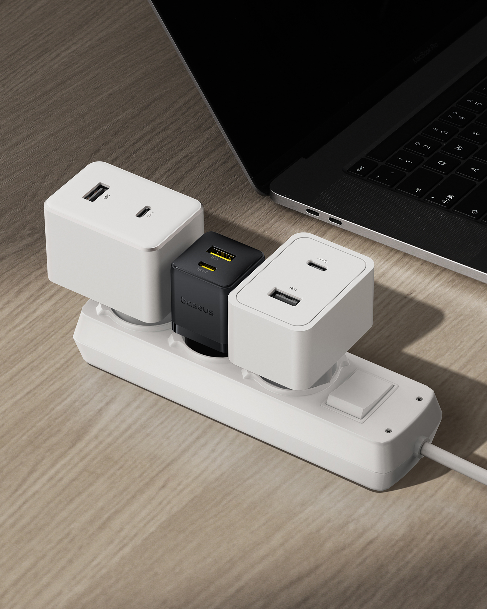 Három hálózati adapter egy fehér elosztóba dugva egy fa felületen, a háttérben egy laptop.