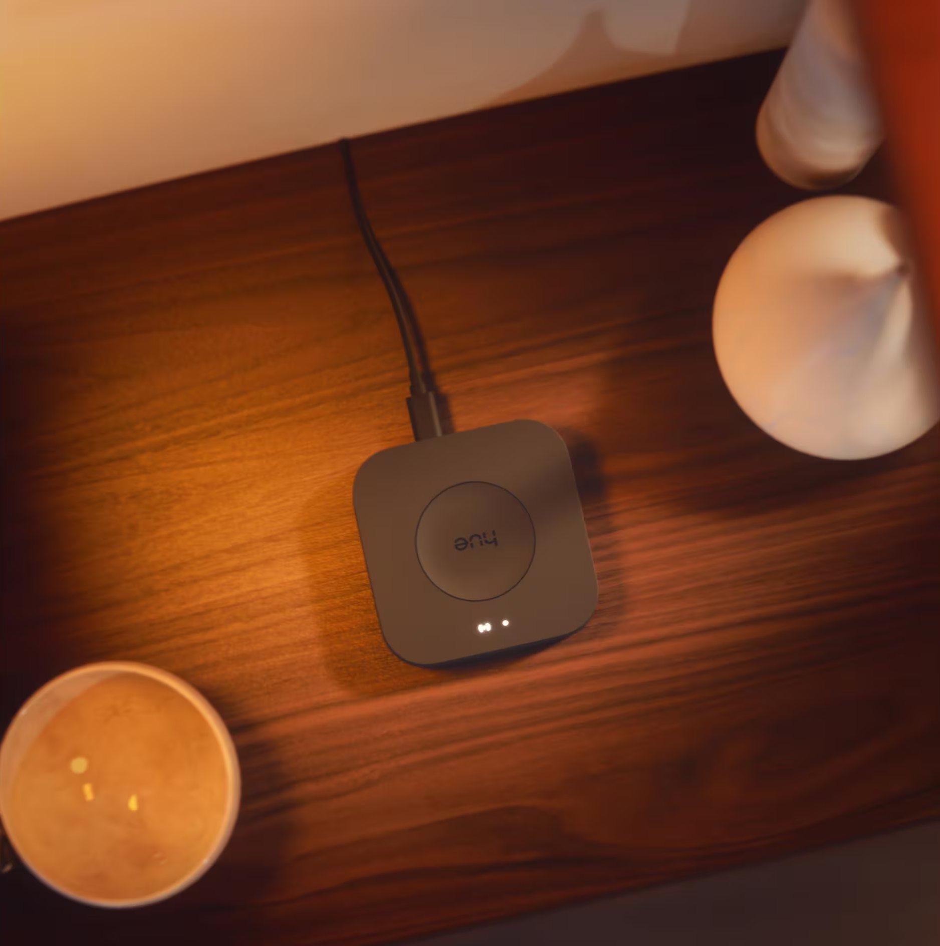 Un appareil Hue noir avec un câble sur une surface en bois, à côté d'une tasse et d'objets blancs.