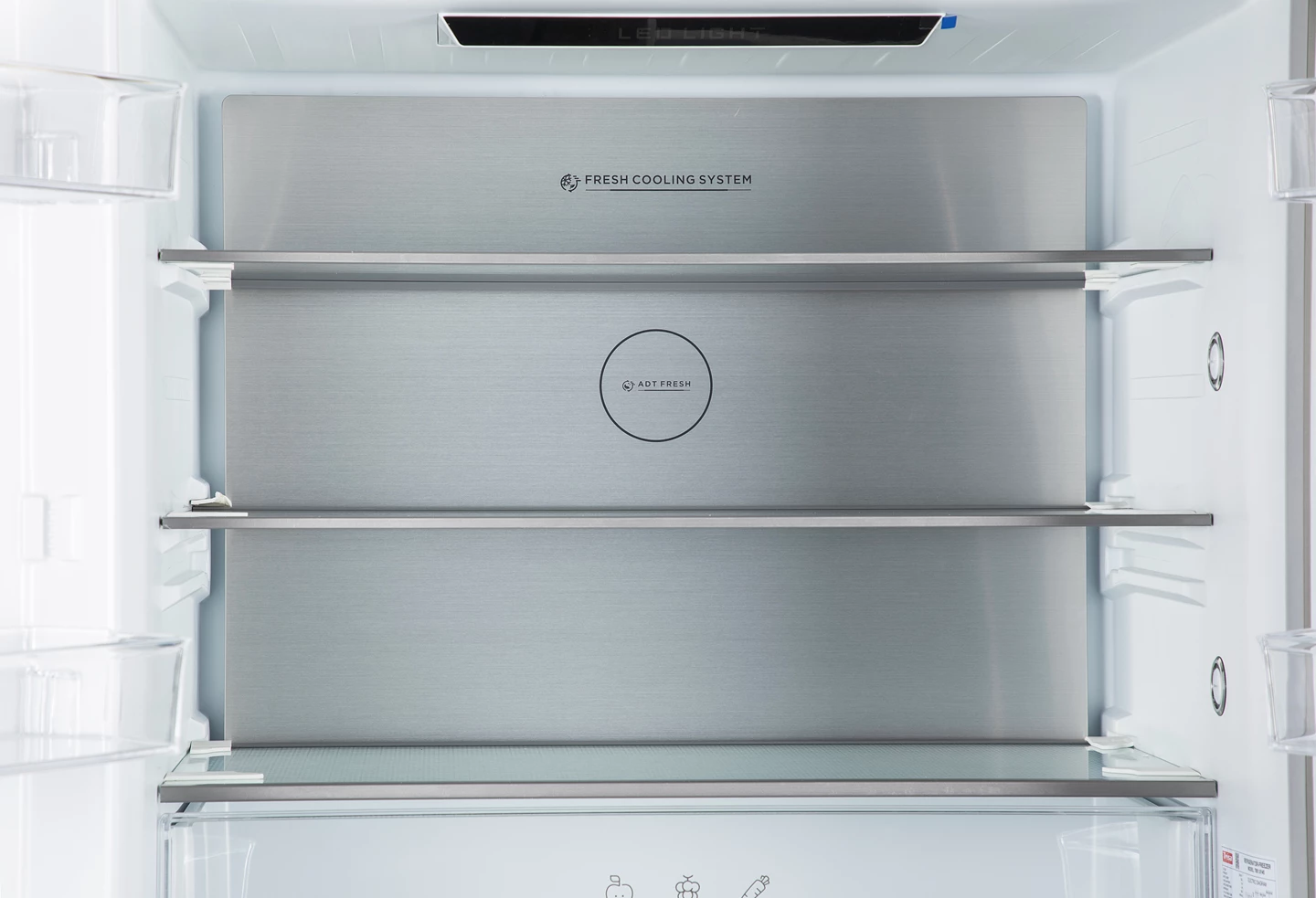Innenansicht eines Kühlschranks mit Regalen. Eine Edelstahl-Rückwand hat Text: FRESH COOLING SYSTEM und ADT FRESH. LED-Licht oben.
