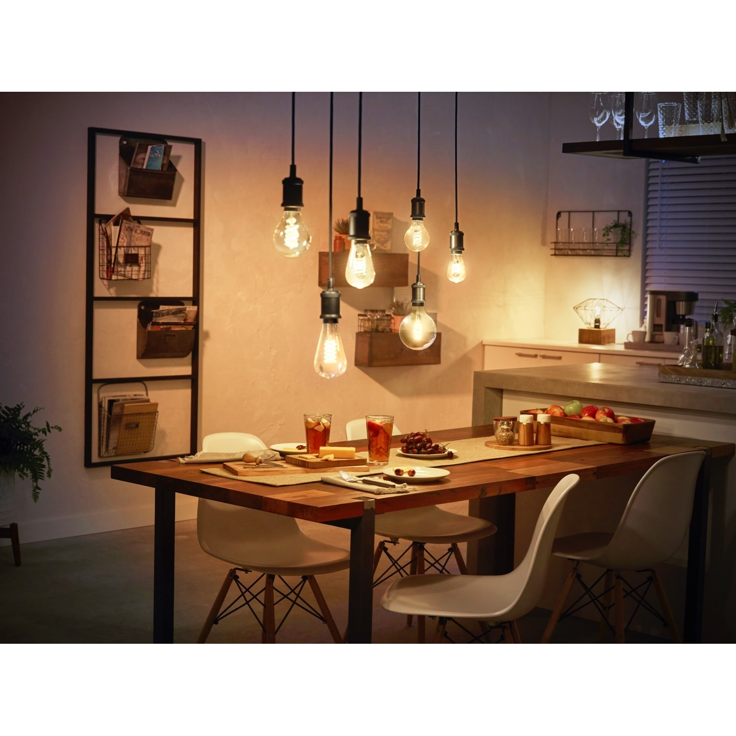 Sala da pranzo con tavolo in legno, sedie bianche, luci a sospensione. Bevande e cibo sul tavolo. Cucina sullo sfondo.