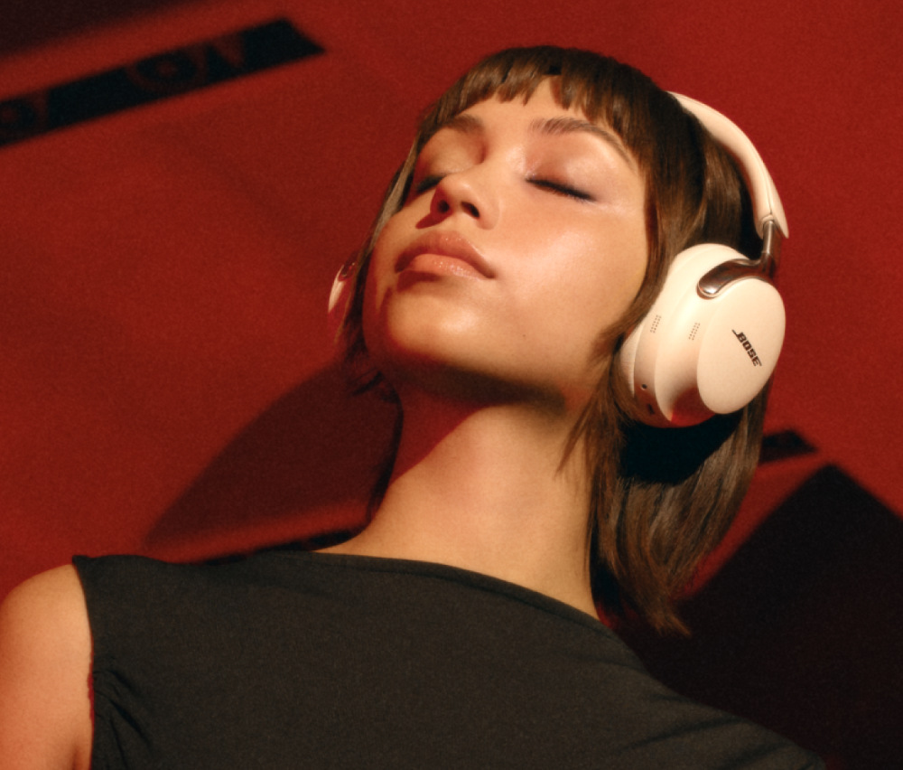 Kobieta z zamkniętymi oczami w białych słuchawkach na czerwonym tle. Ma na sobie czarny top. Słuchawki marki Bose.