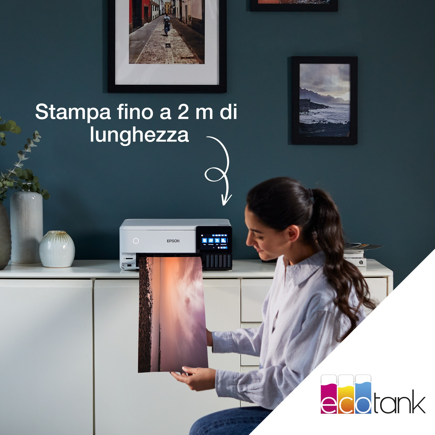 Una donna tiene in mano una stampa di grandi dimensioni da una stampante Epson. La stampa ha una foto del tramonto. Lo sfondo ha quadri con cornice.