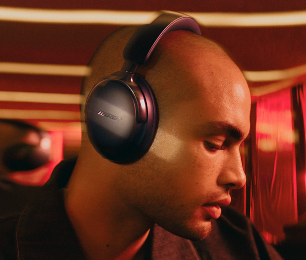 Łysy mężczyzna w czarnych słuchawkach Bose. Tło jest czerwone z paskami i delikatną poświatą.