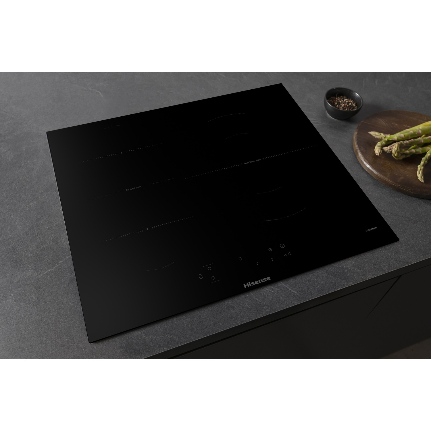 Un piano cottura a induzione Hisense nero su un piano scuro. Una ciotola di spezie e asparagi su un tagliere di legno.