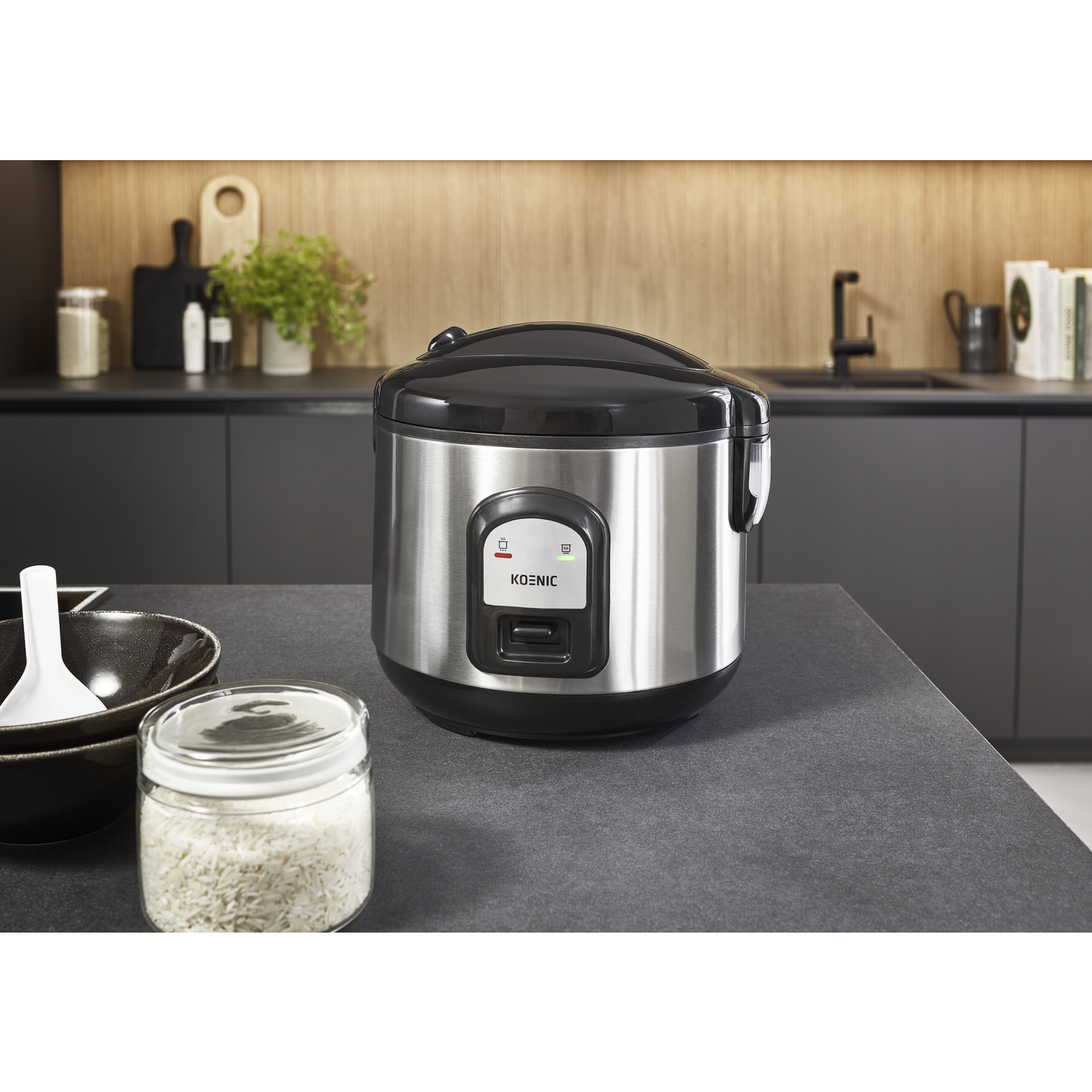 A stainless steel rice cooker sits on a countertop with a jar of rice and bowls. The background shows a kitchen setting.