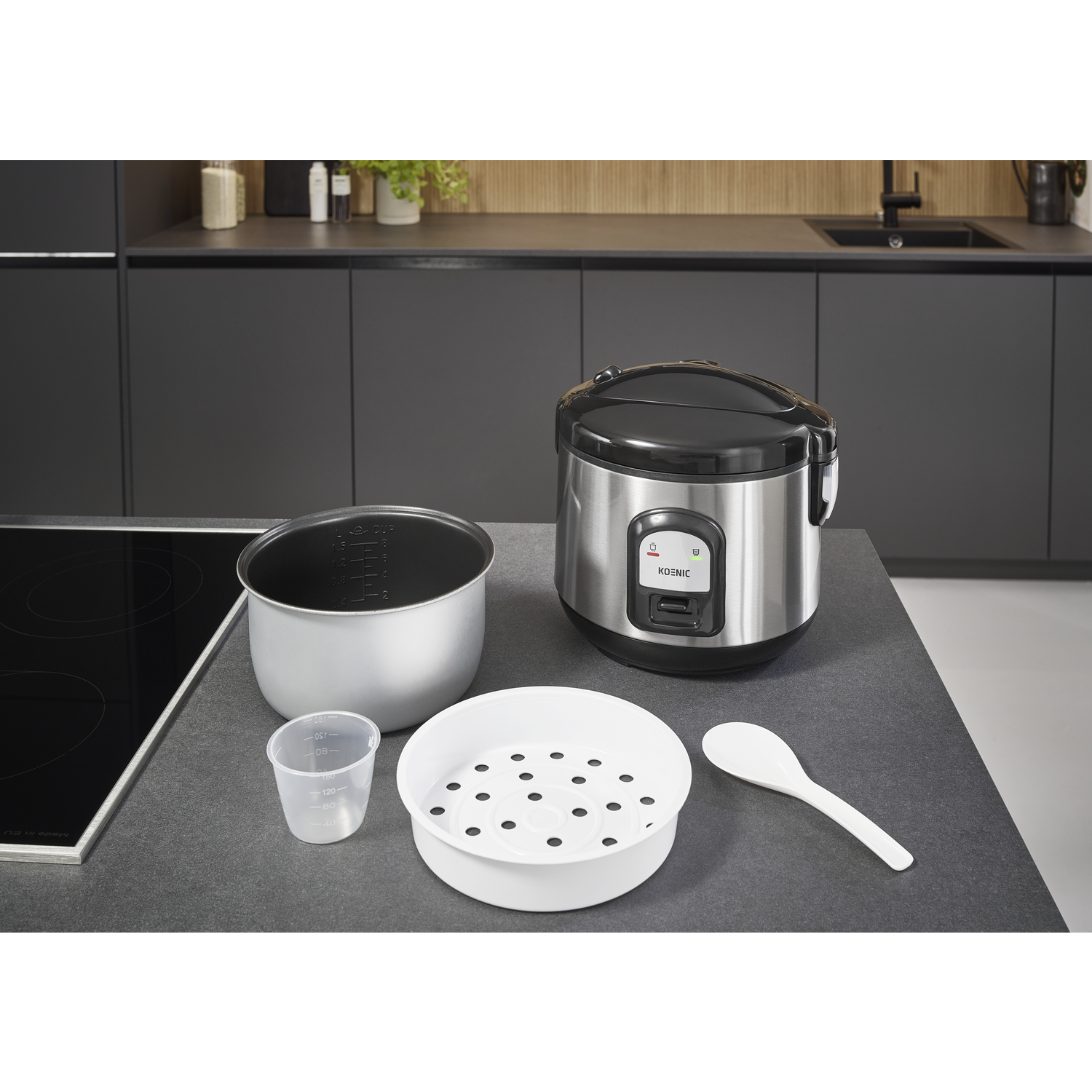 A stainless steel rice cooker with accessories: inner pot, measuring cup, steamer, and spoon. The cooker is on a dark countertop.