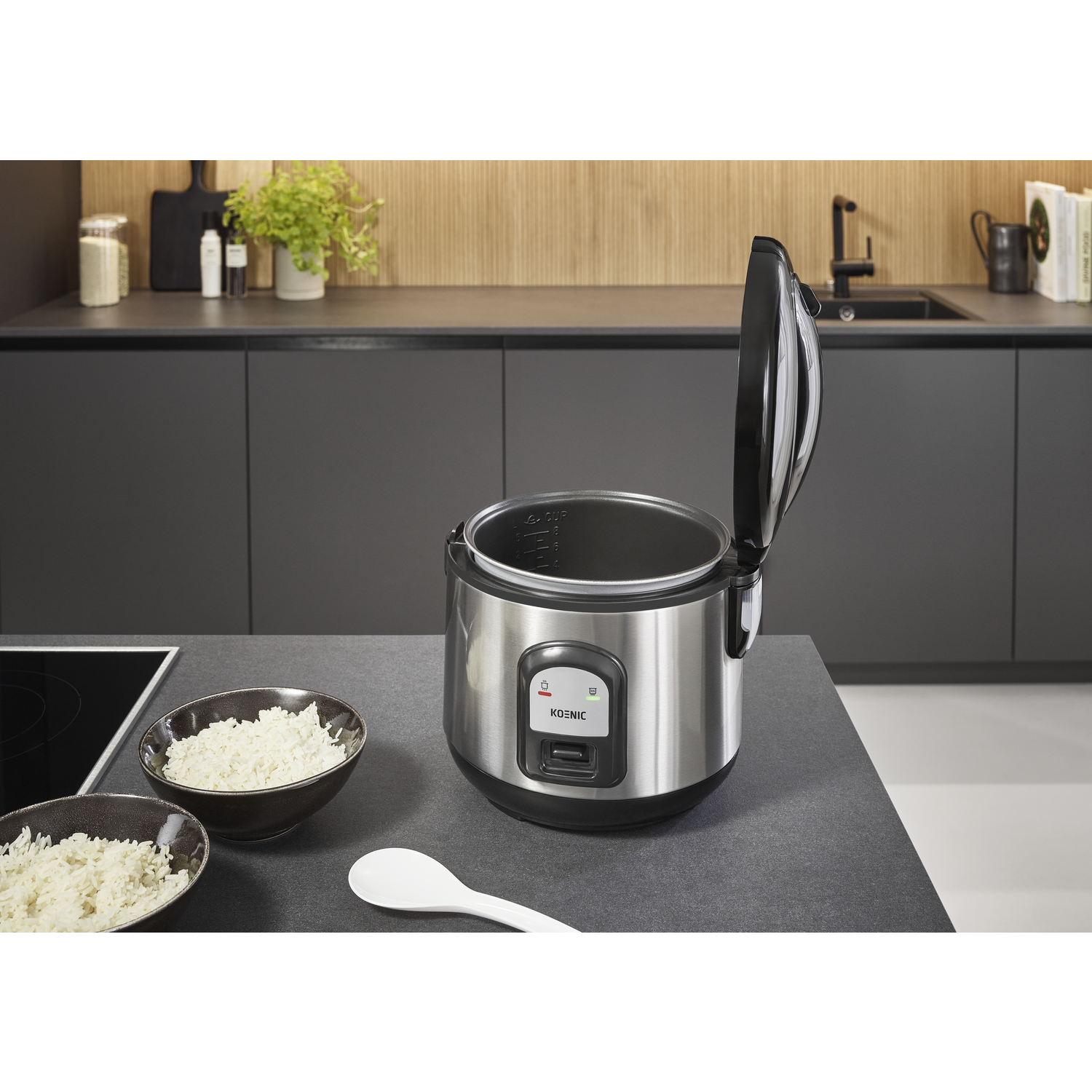 A stainless steel rice cooker with an open lid stands on a countertop. Two bowls of rice and a white spoon are in front.