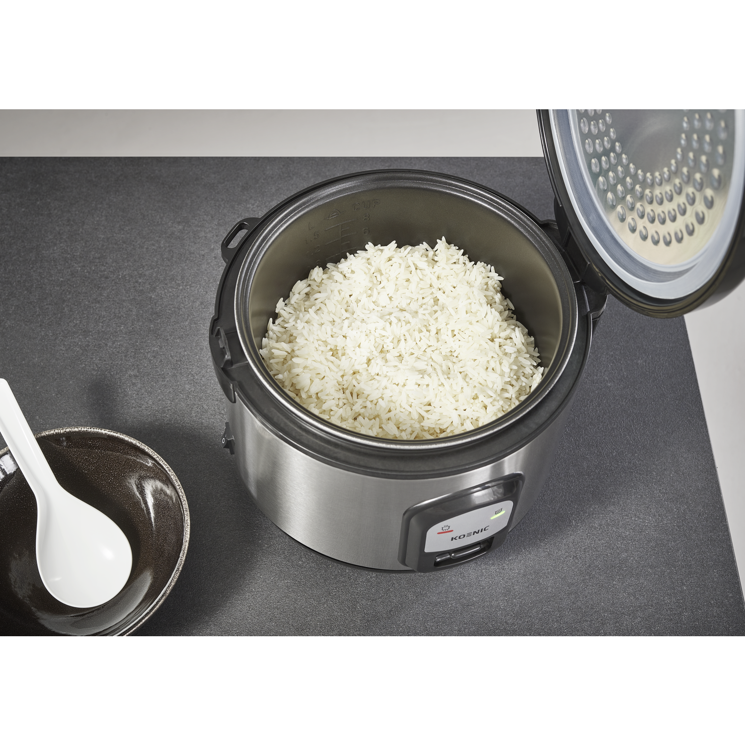 Rice cooker with cooked white rice, open lid. A bowl with a white spoon is on the left. Grey countertop.