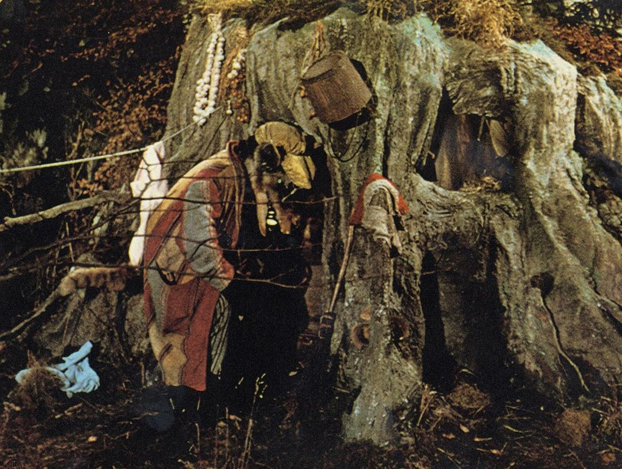 Eine kostümierte Figur steht in der Nähe eines großen Baumstamms. Ein Eimer hängt darüber. In der Nähe befinden sich ein Mopp und eine Stoffpuppe. Der Hintergrund ist dunkel.