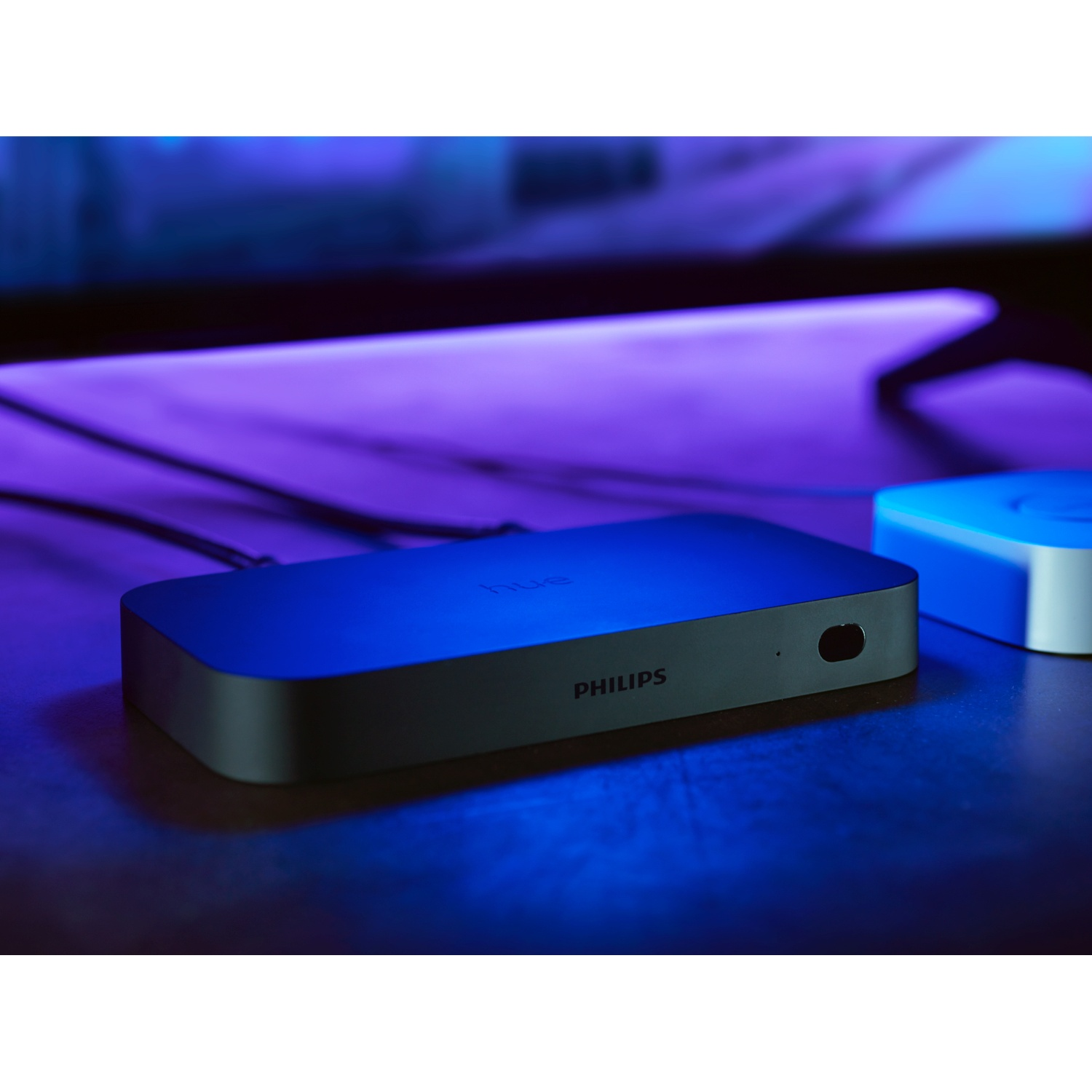 Un bridge Philips Hue nero è su un tavolo. La parola 'HUE' è visibile. Una luce blu lo circonda.