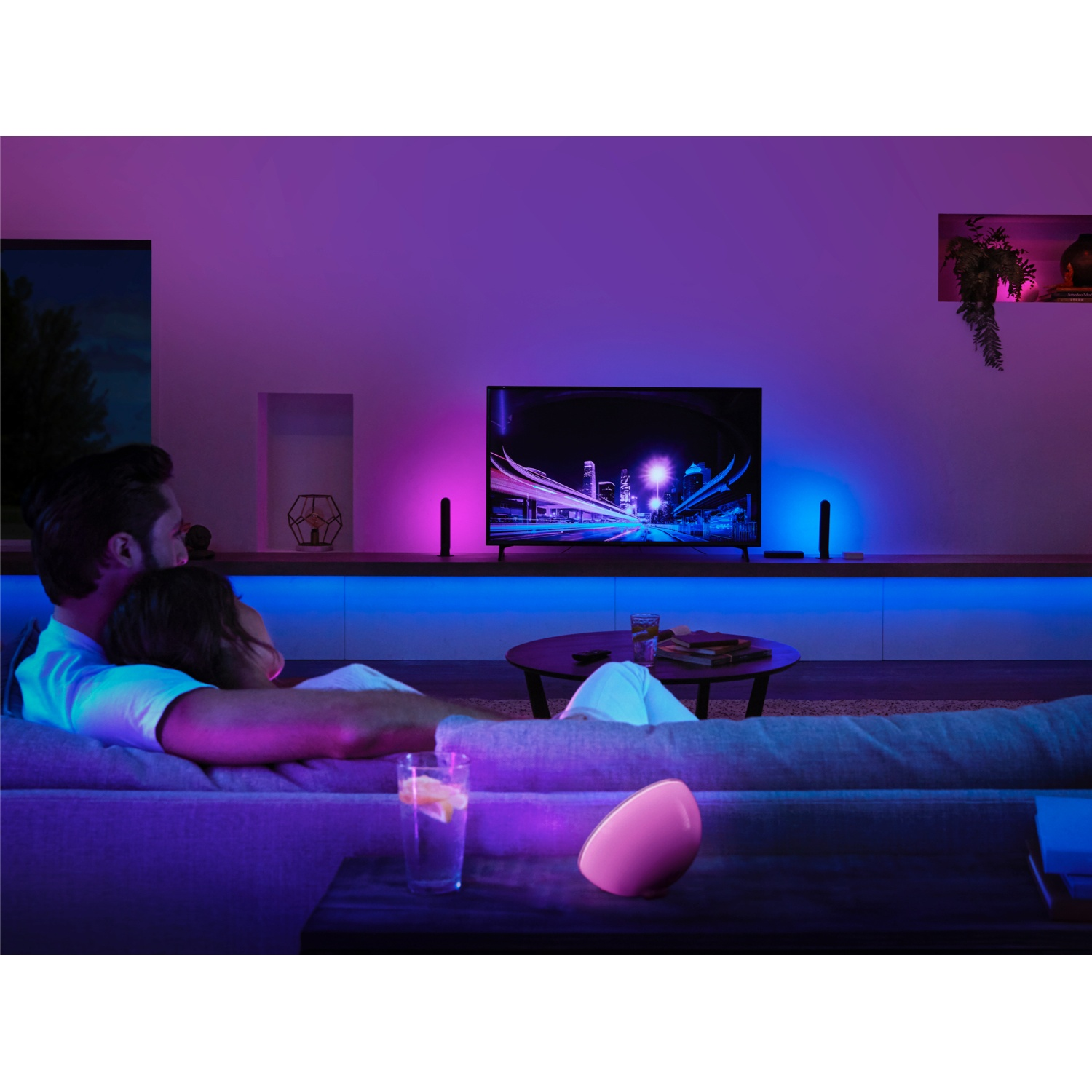 Coppia sul divano guarda la TV con luci della città. Lampada rosa sul tavolo. Illuminazione ambientale blu e viola.