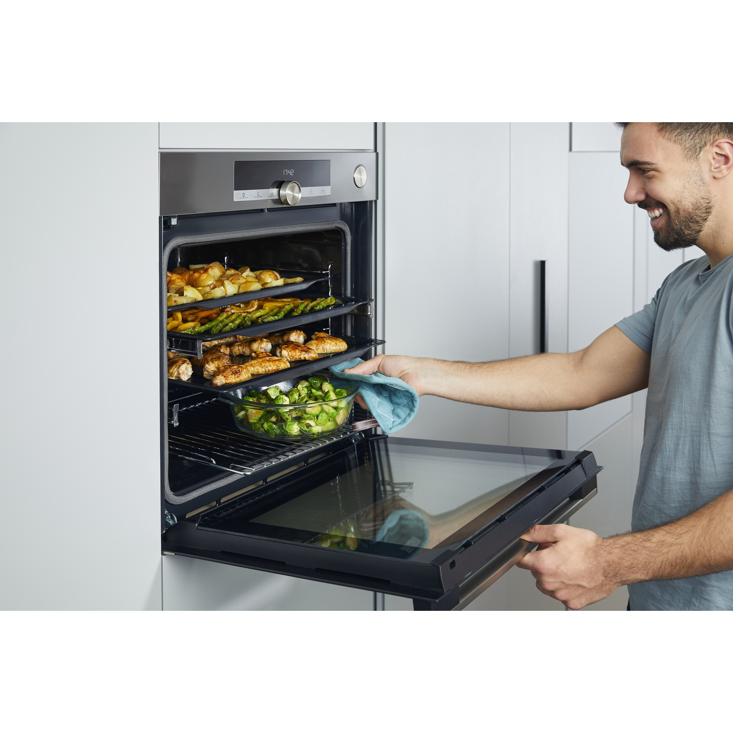 Un uomo sorride aprendo la porta del forno. All'interno ci sono vassoi di cibo cotto: patate, asparagi, pollo e cavolini di Bruxelles.