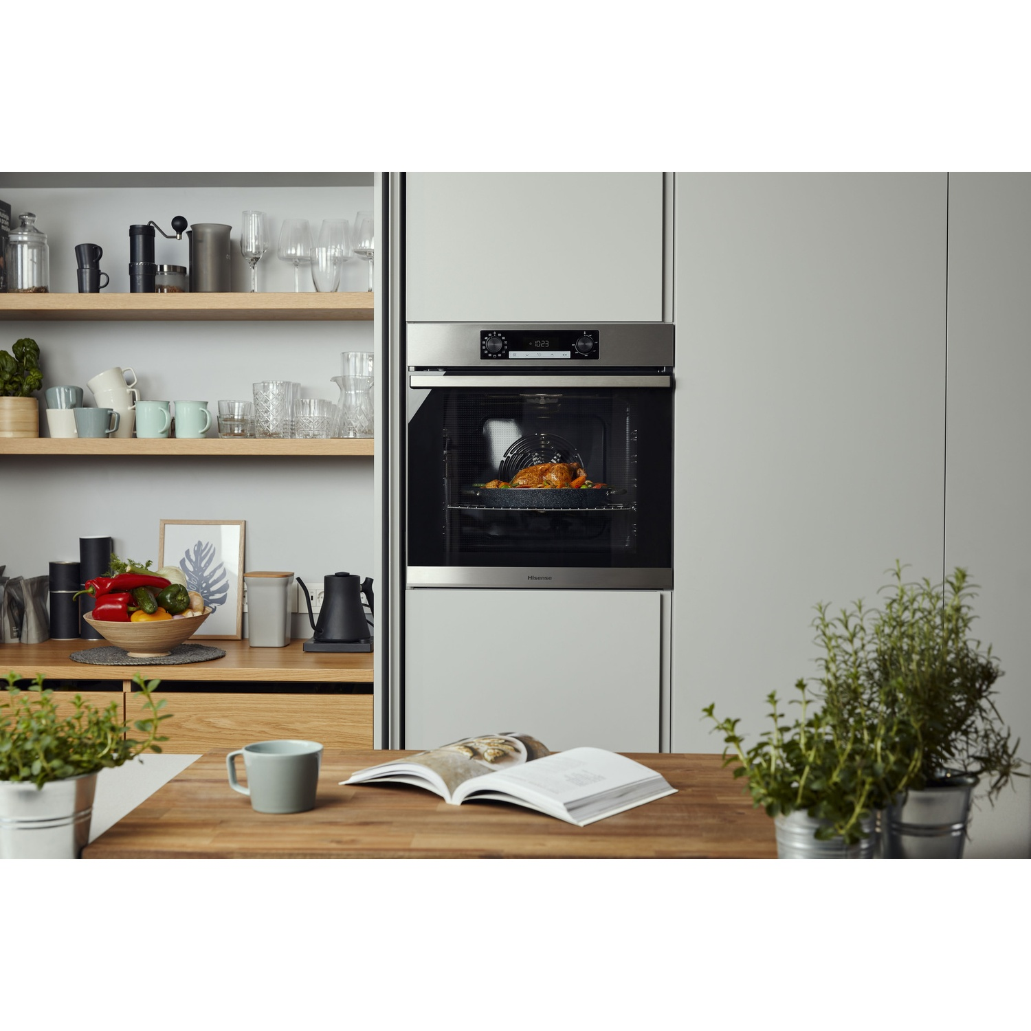 Una scena di cucina con un forno contenente un pollo arrosto. Un tavolo di legno con tazza, piante e un libro aperto. Scaffali mostrano utensili da cucina.