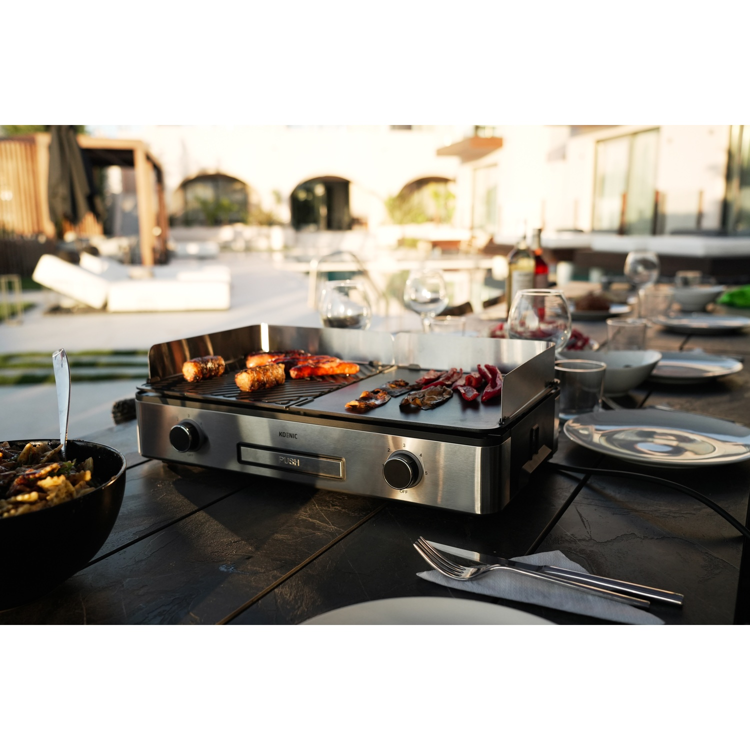 Een grill op een tafel met eten. Borden, glazen en bestek zijn gedekt. De scène is een openluchtdiner met een gebouw op de achtergrond.