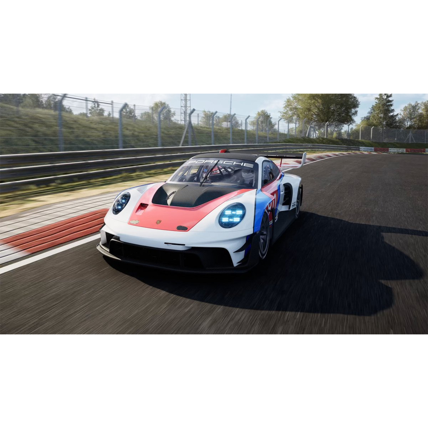 A white, red, and blue Porsche race car speeds around a racetrack. Trees and a fence are in the background under a blue sky.
