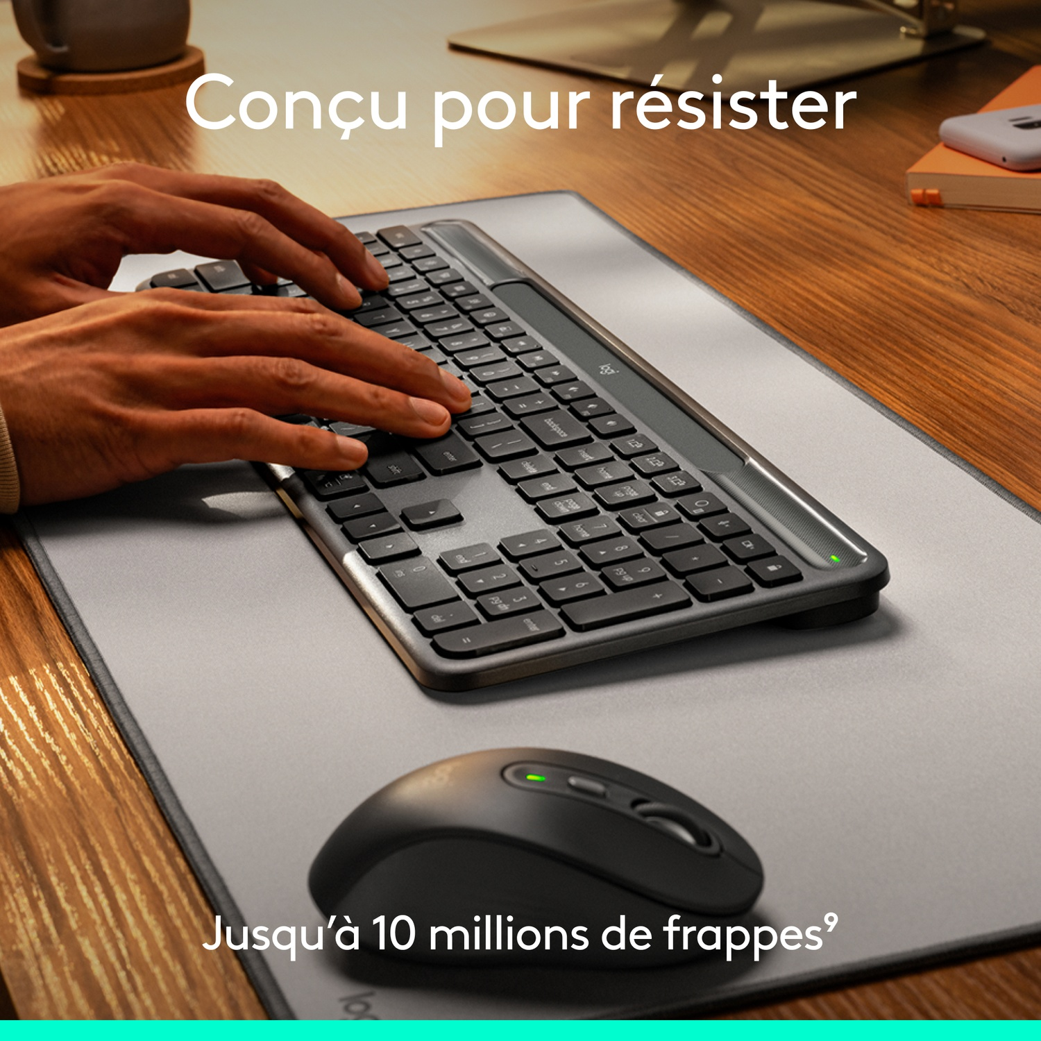 Mains tapant sur un clavier. Une souris noire est en dessous. Le clavier et la souris sont sur un tapis gris, sur un bureau en bois.