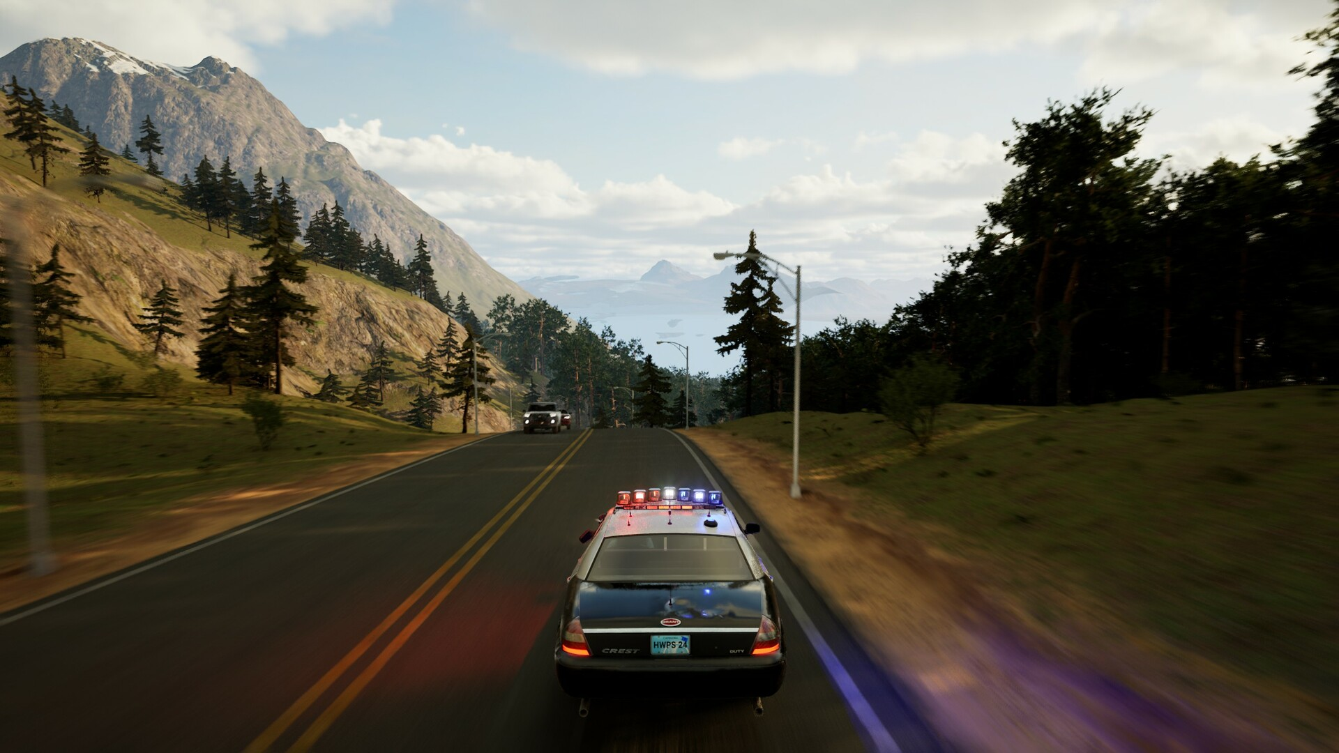 Une voiture de police roule sur une route, des montagnes et des arbres en arrière-plan. Une autre voiture de police est devant. Ciel bleu avec des nuages.