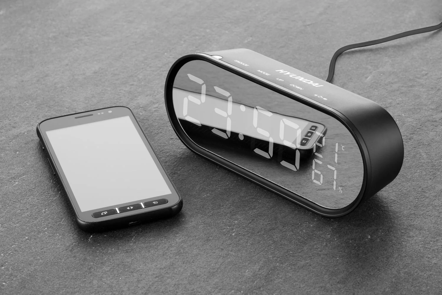 Czarny zegar cyfrowy z telefonem obok. Zegar pokazuje godzinę 23:50 oraz temperaturę i wilgotność. Szare tło.