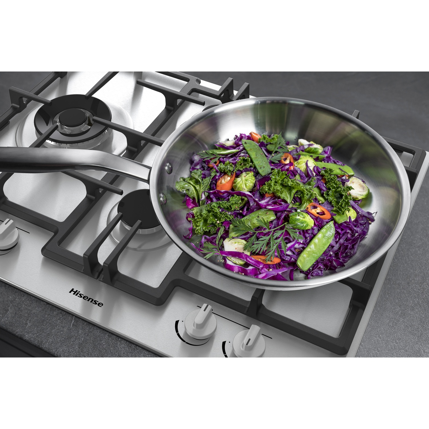 Una padella in acciaio inox con verdure colorate si trova su un piano cottura a gas. Le verdure includono cavolo rosso, piselli e peperoni.