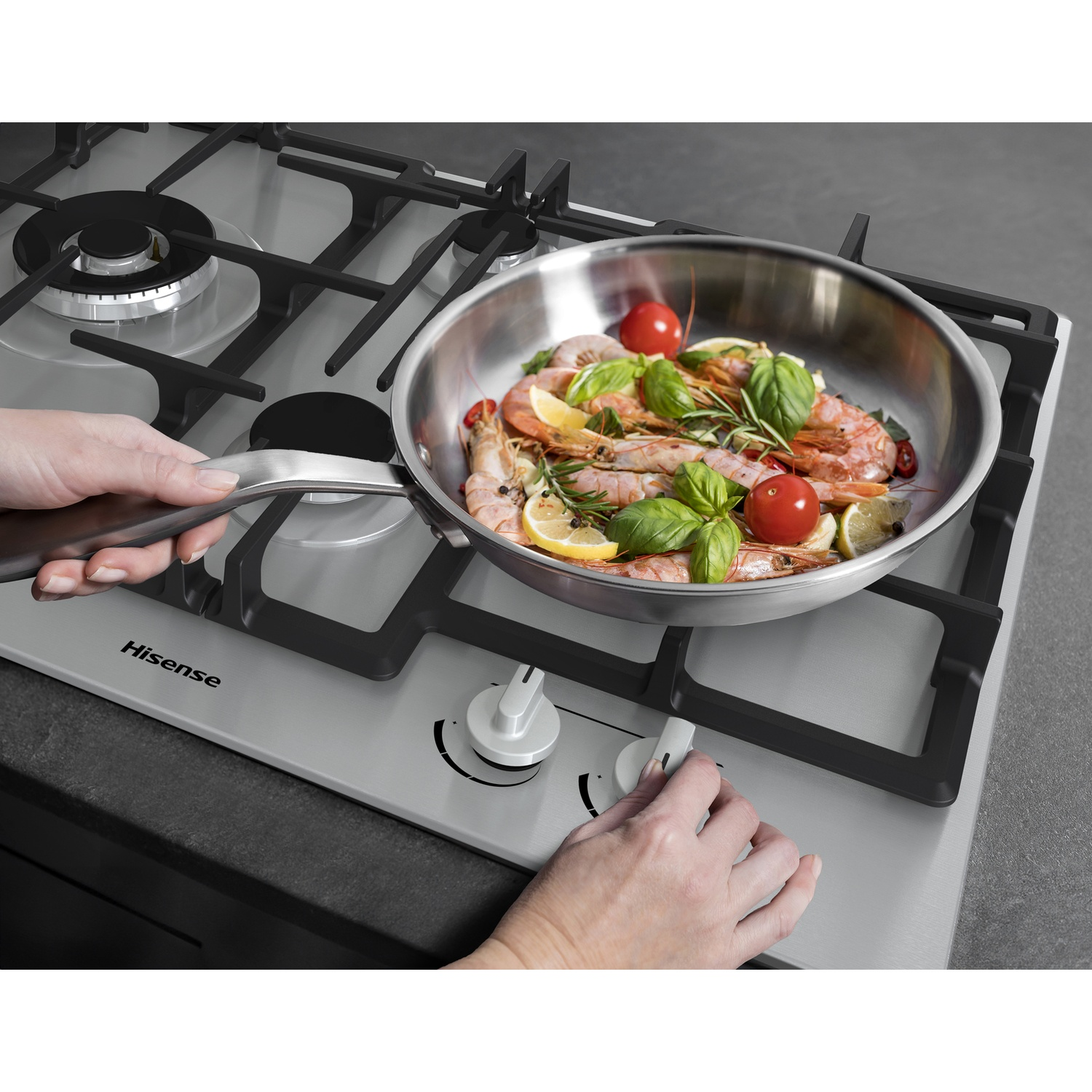 Una padella in acciaio inox con gamberi, pomodori, basilico e limone su un fornello. Mani che regolano le manopole del fornello.