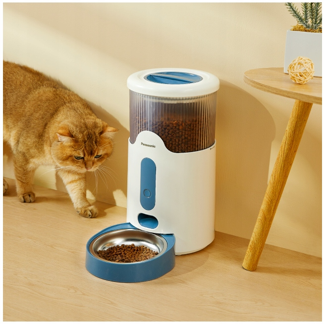 Kot zbliża się do białego i niebieskiego automatycznego podajnika karmy. Podajnik ma miskę z karmą. Po prawej stronie znajduje się stolik pomocniczy.