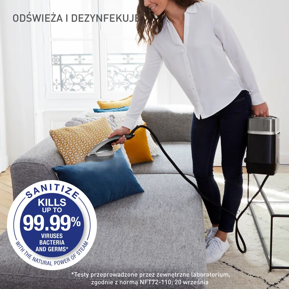 Kobieta paruje kanapę czarnym parowarem. Kanapa ma kolorowe poduszki. Tekst mówi 'dezynfekcja zabija do 99,99% wirusów'.