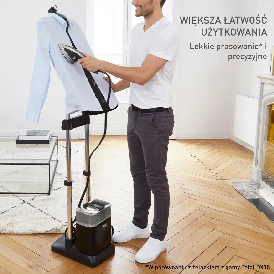 Mężczyzna prasuje niebieską koszulę na parownicy do ubrań. Ma na sobie białe tenisówki, białą koszulę i szare spodnie. Tło to pokój.