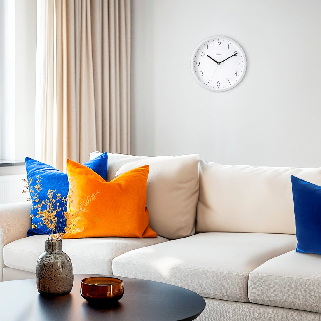 Biały zegar ścienny wisi nad białą sofą z niebieskimi i pomarańczowymi poduszkami. Stół zawiera wazon i małą miskę.