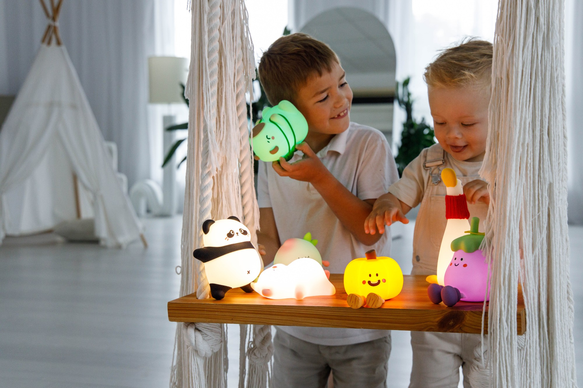 Dwoje dzieci bawi się kolorowymi, świecącymi lampami z kreskówek na drewnianej huśtawce. Tło obejmuje tipi i białą zasłonę.