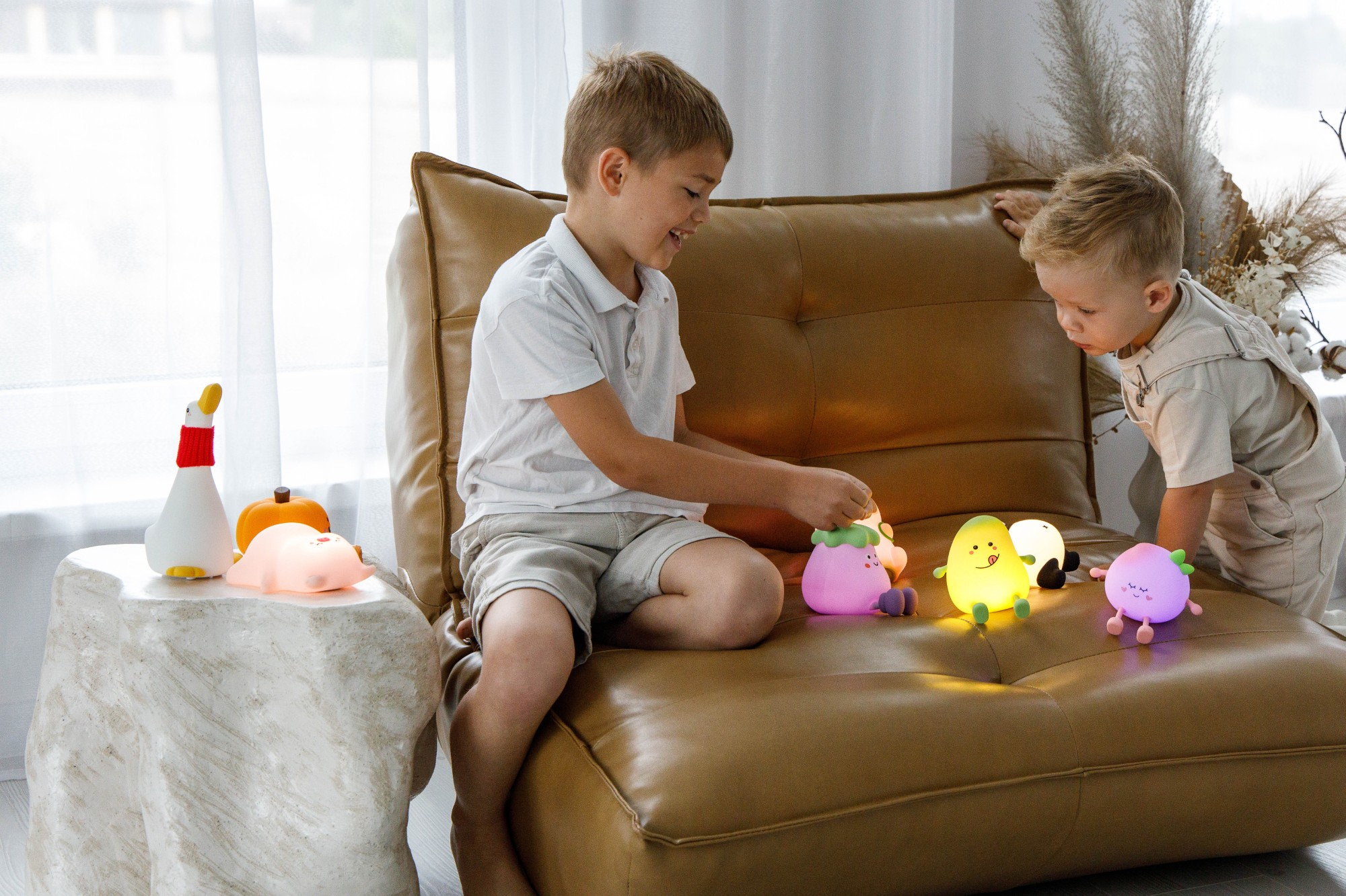 Dwóch chłopców bawi się świecącymi lampkami w kształcie owoców. Na białym stoliku lampa w kształcie kaczki i dyni. Brązowa kanapa i białe zasłony.