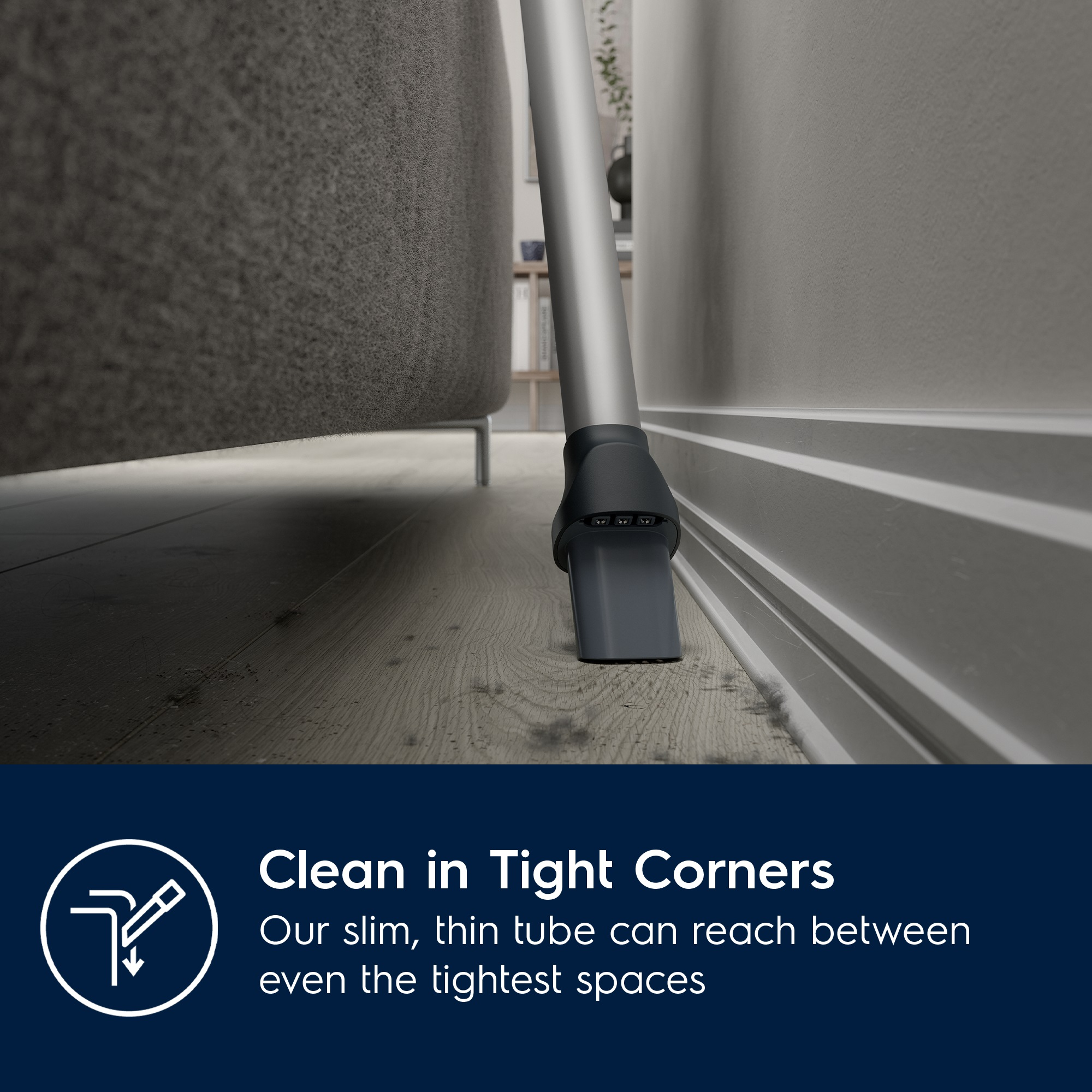 Bir elektrik süpürgesinin başlığı bir duvarın ve bir kanepenin yakınındadır. Zemin ahşaptır. Metin şöyle diyor: 'Clean in Tight Corners'.
