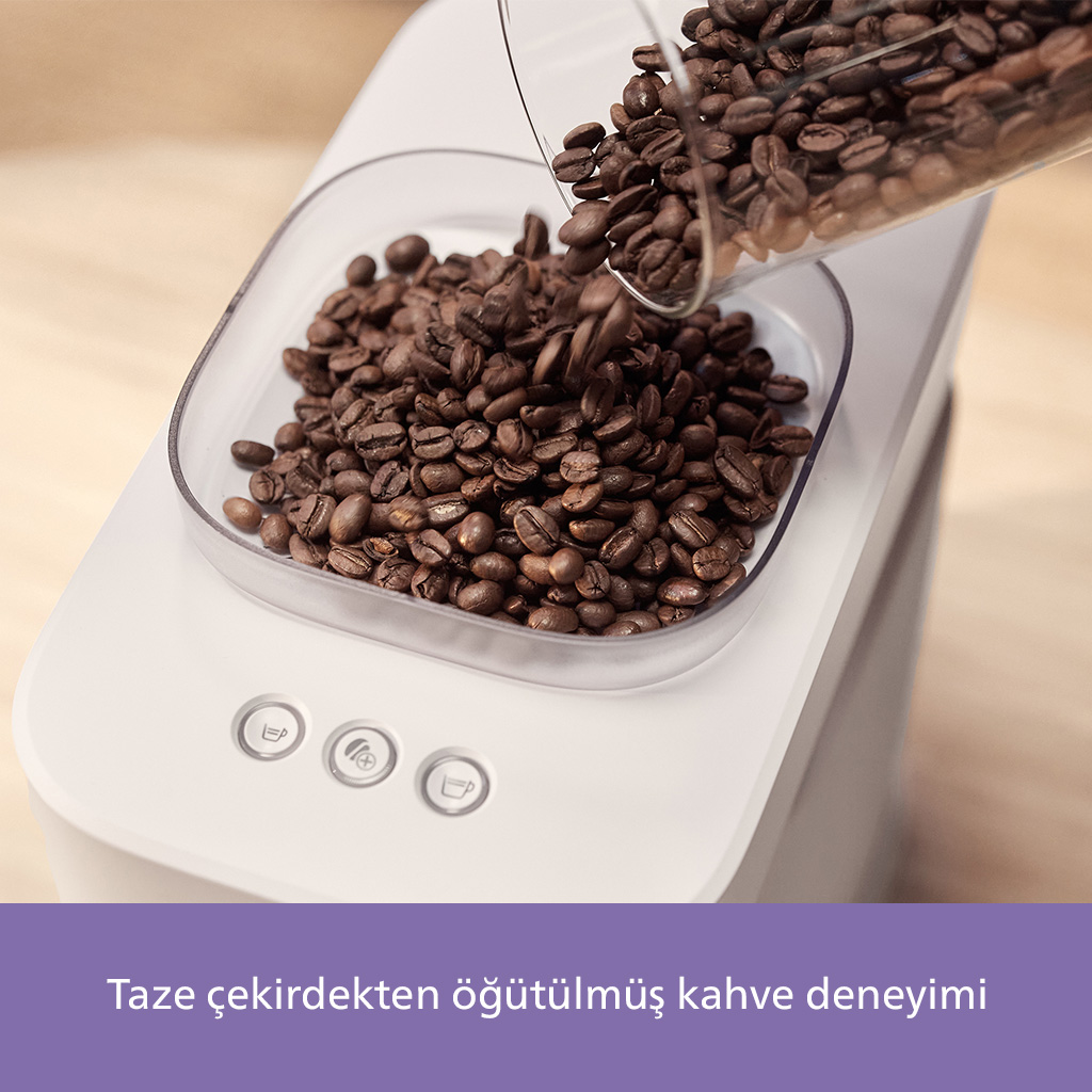 Kahve çekirdekleri, beyaz bir kahve makinesinin üzerindeki şeffaf bir kaba dökülüyor. Üç düğme görünür. Arka planda ahşap bir yüzey.