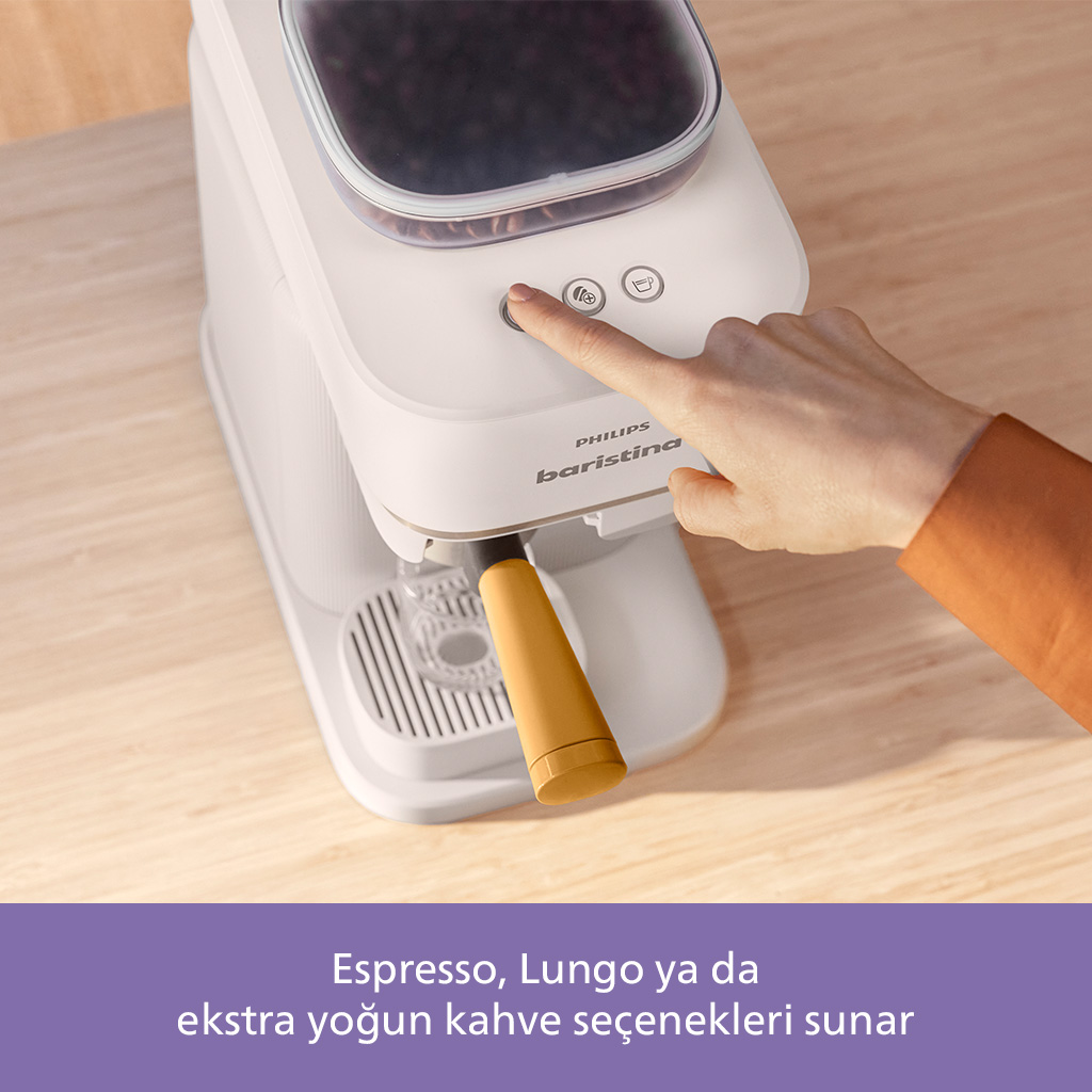Bir el, beyaz bir Philips barista makinesindeki bir düğmeye basıyor. Bir kapta kahve çekirdekleri. Sap sarı.