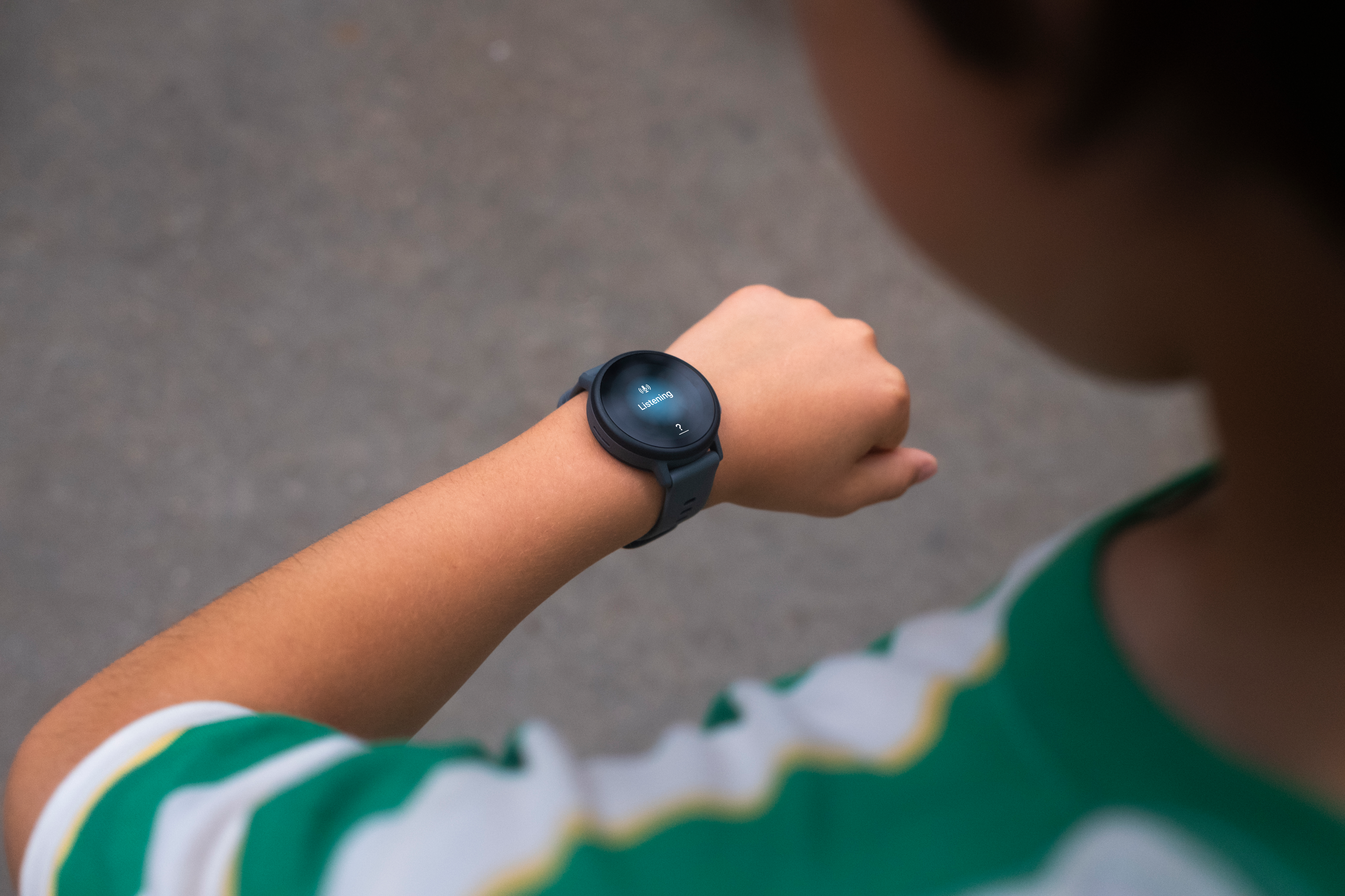 Eine Person trägt eine Smartwatch, das Ziffernblatt zeigt 'Listening'. Die Person trägt ein grün-weiß gestreiftes Hemd.