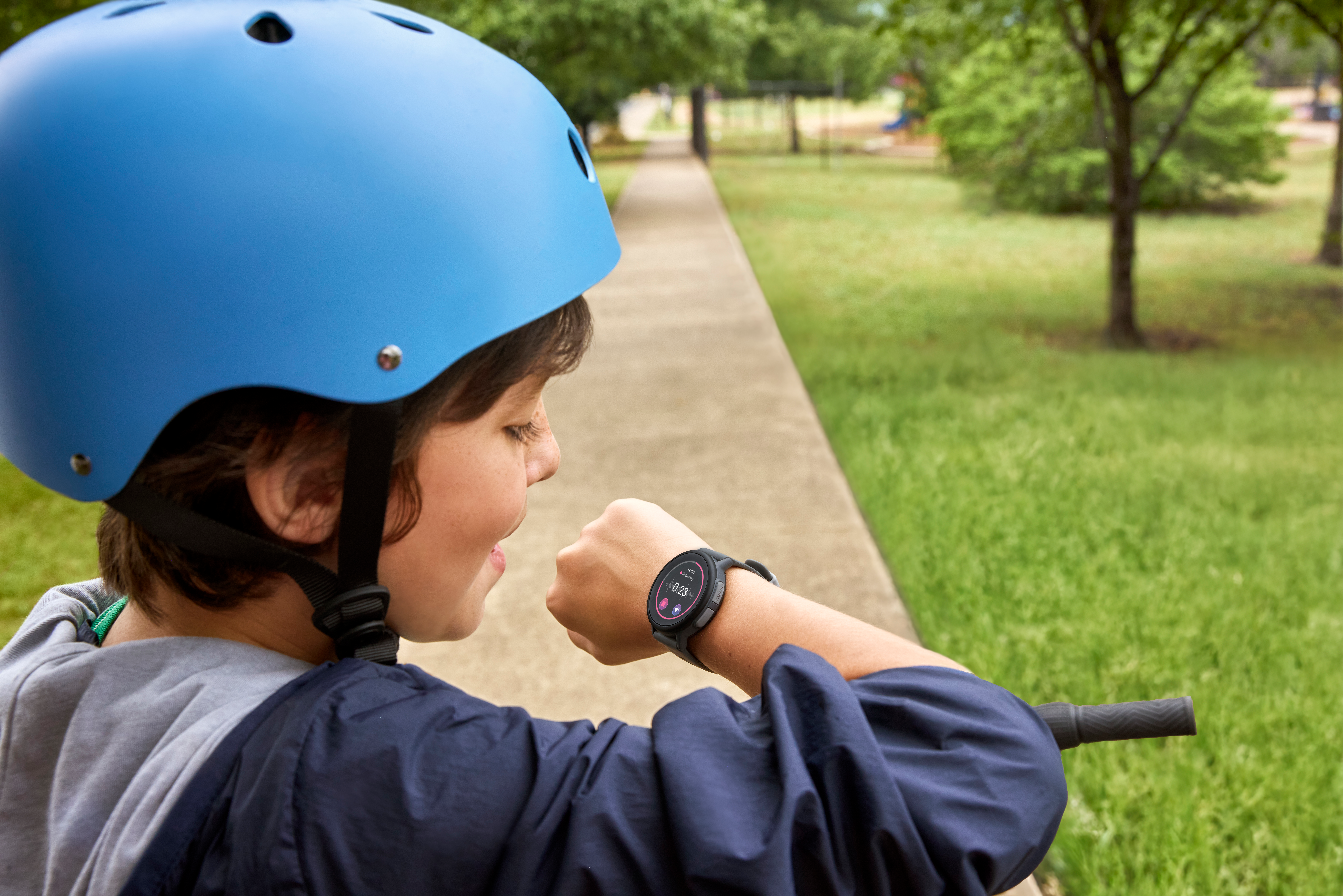 Ein Kind mit blauem Helm betrachtet eine Smartwatch am Handgelenk. Im Hintergrund sind ein Weg und grünes Gras zu sehen.